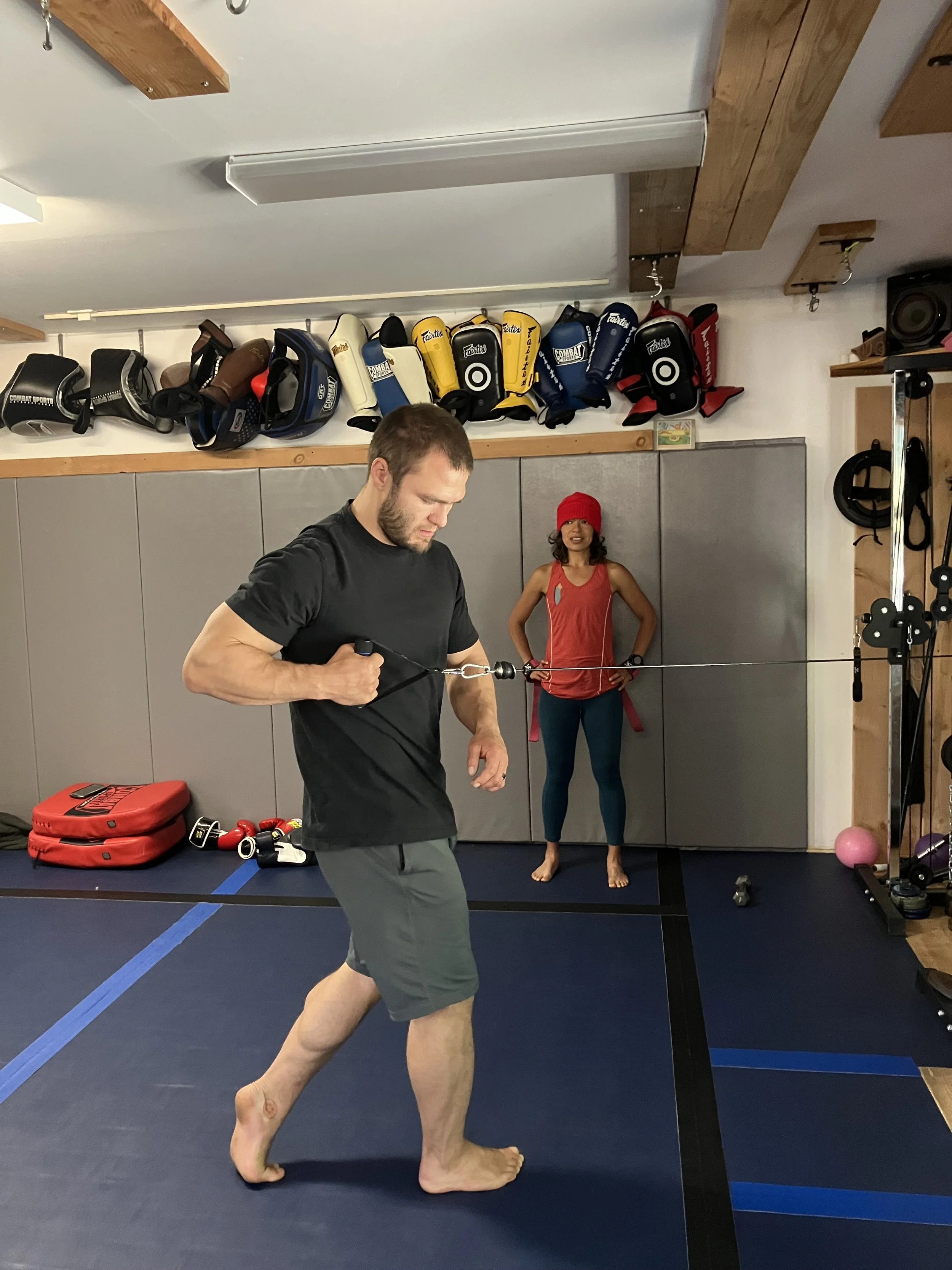 Man practicing boxing with a resistance band in a gym, woman watching in the background, boxing gloves and equipment on shelves