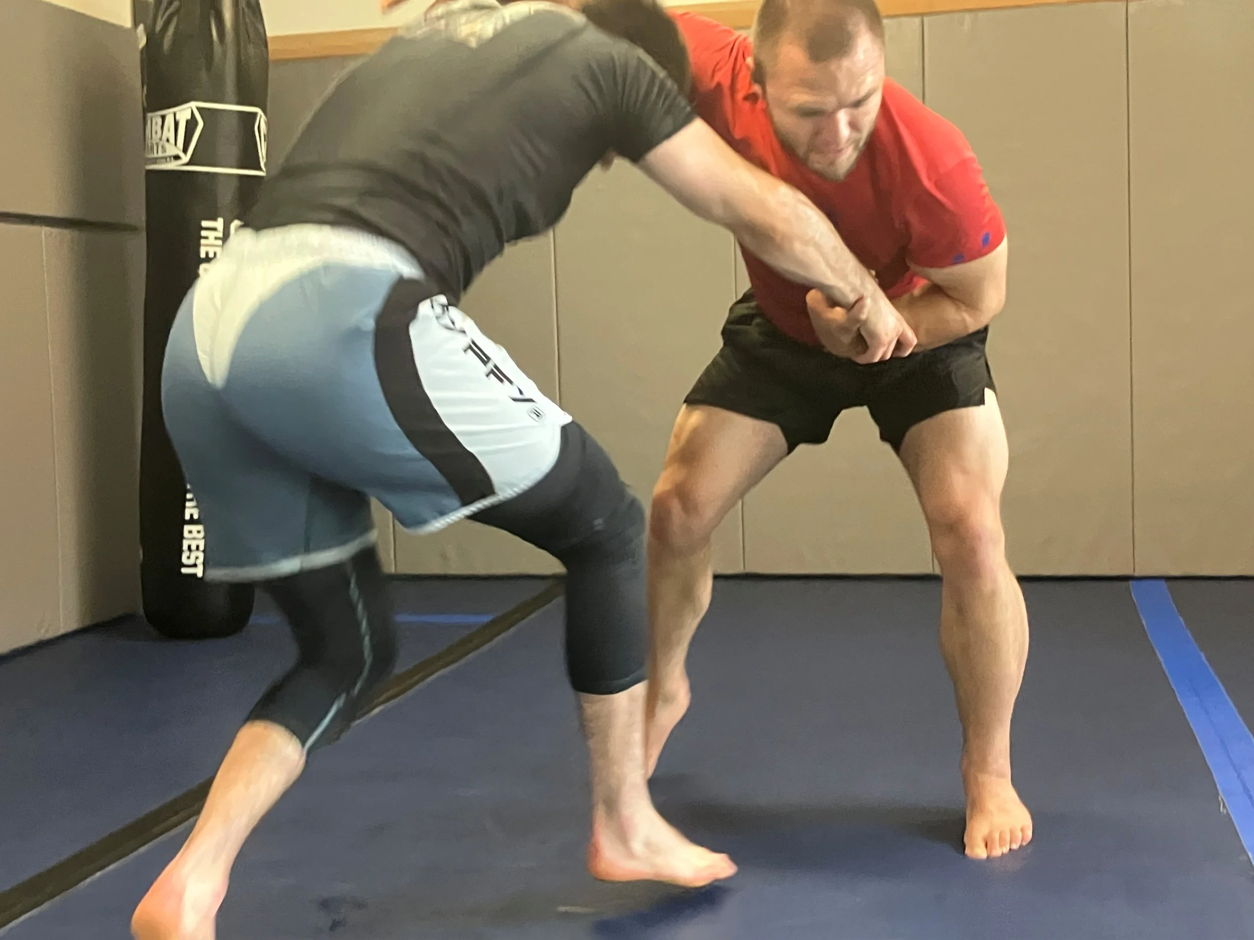 Two men sparring in mixed martial arts inside a gym. One man in black compression pants and a black T-shirt is grappling the other man in red shirt and black shorts. They are on a blue mat near a punching bag in the background.