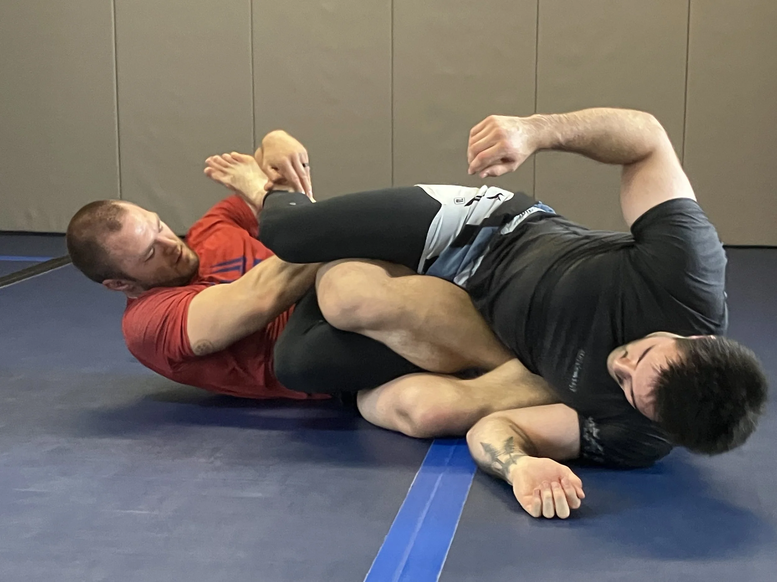 Two men grappling on a blue mat, practicing Brazilian Jiu-Jitsu. One man in a red shirt is on top, controlling the man in black who is on his side. The scene is indoors with grey walls.