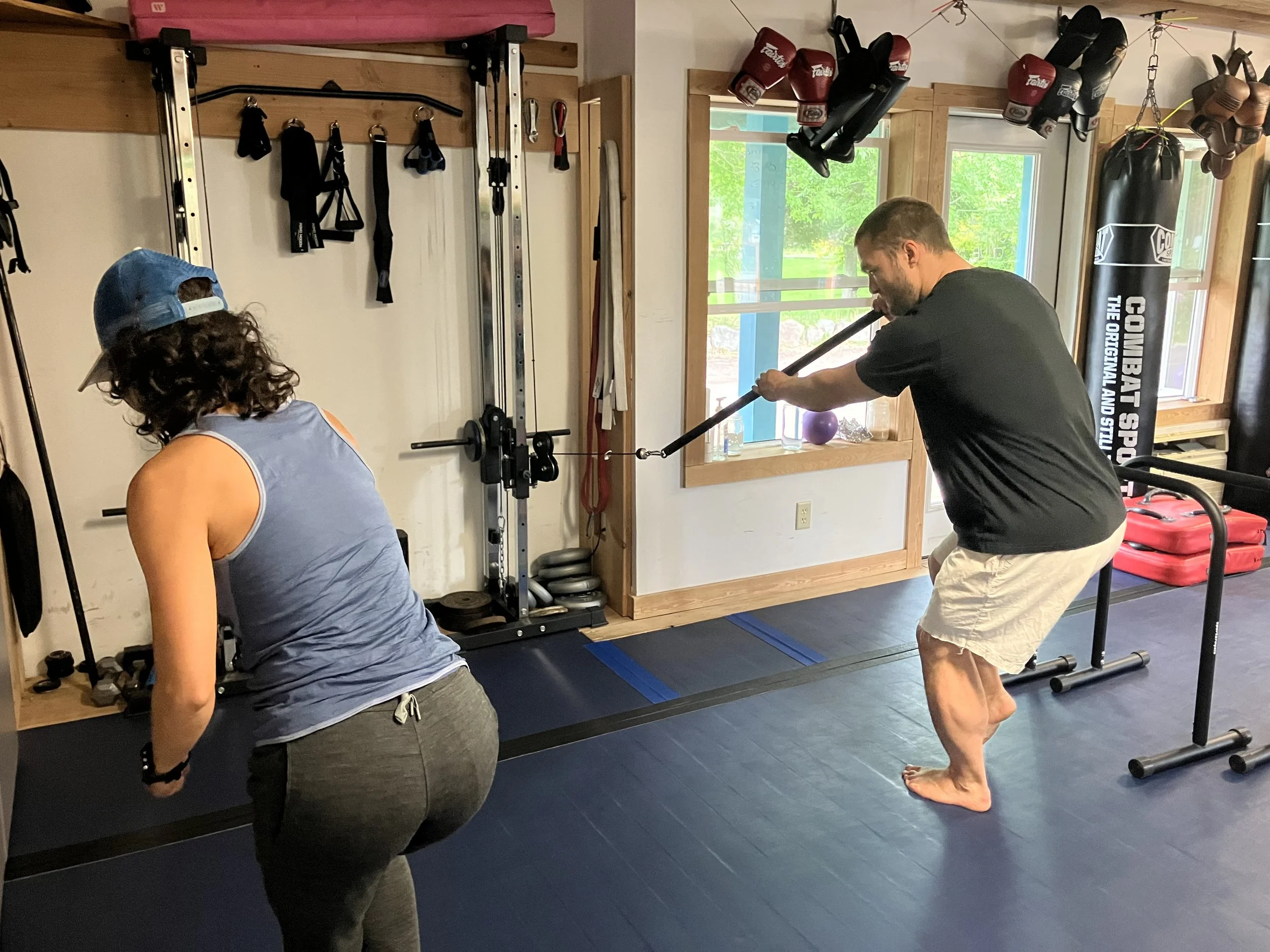 A man and a woman working out in a gym. The man is using a resistance band for exercises, while the woman observes. There are boxing gloves, punching bags, and workout equipment around them.