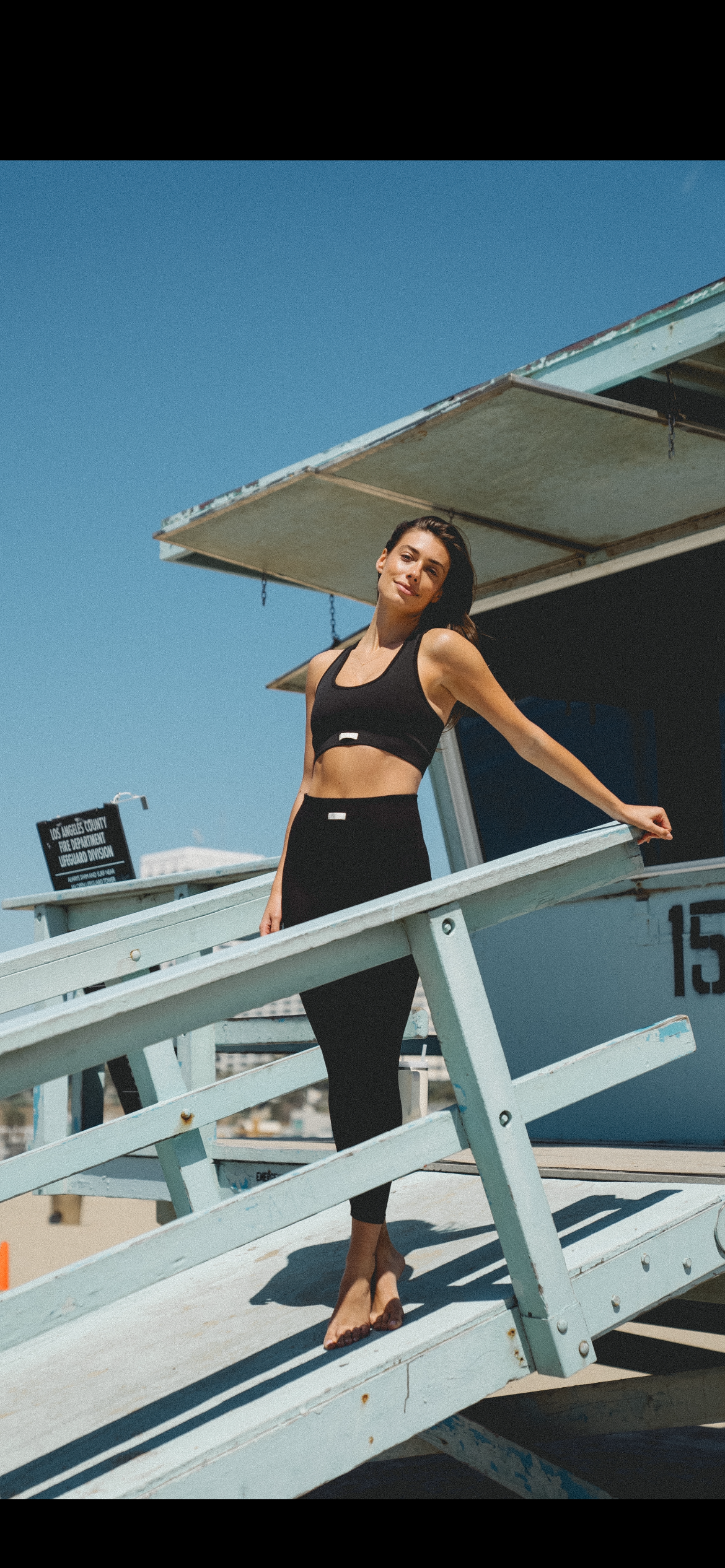A woman in black workout attire standing barefoot on a lifeguard tower at the beach on a sunny day.