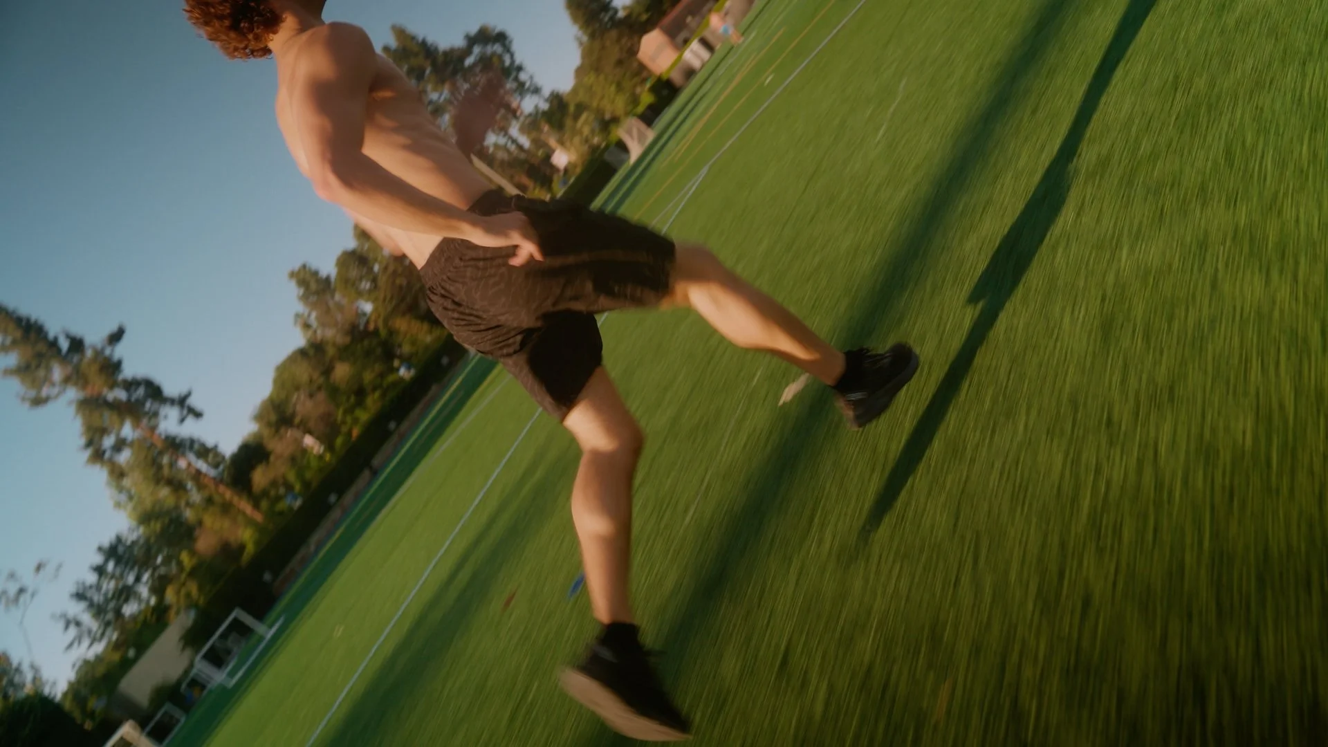 A shirtless man with curly hair running on a green field during sunset.