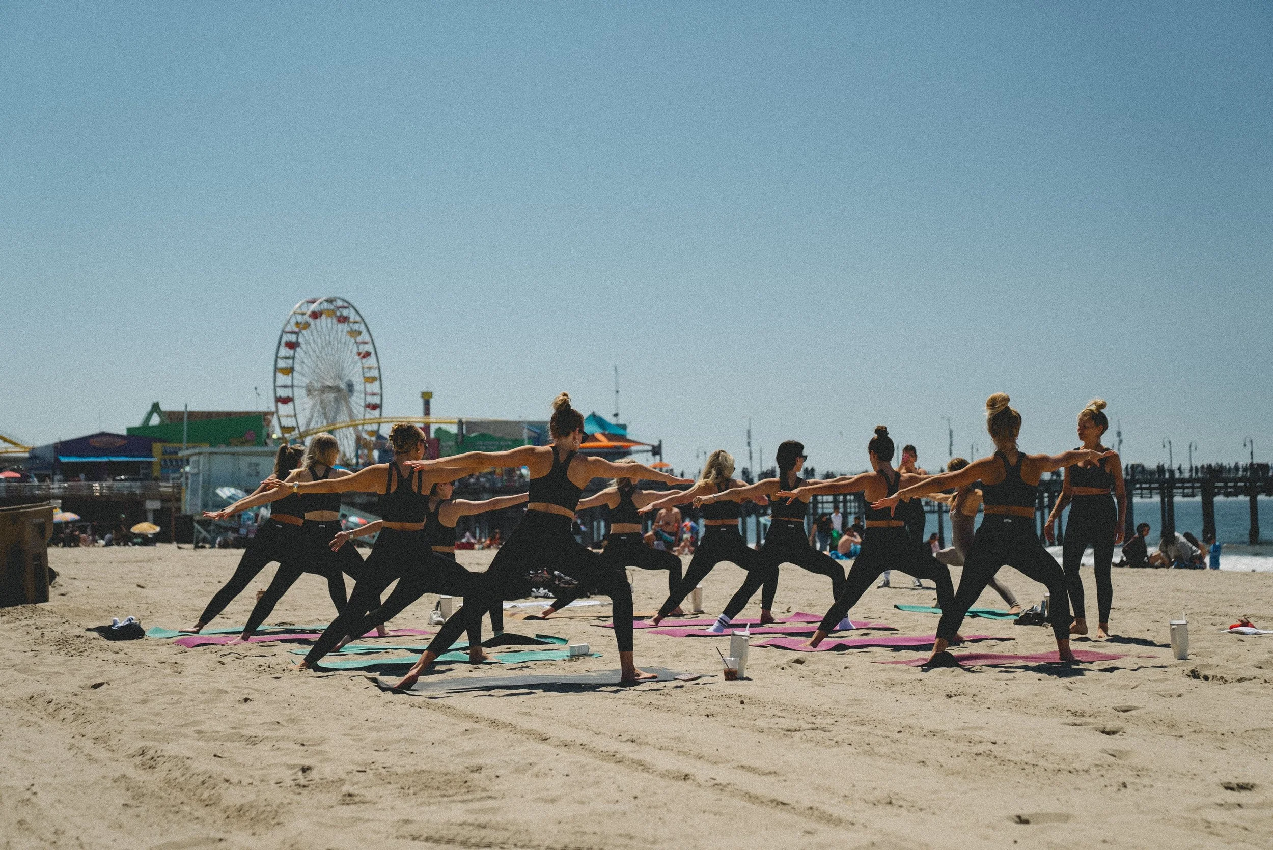 A group of women practicing yoga on the beach with a pier and amusement park rides in the background.