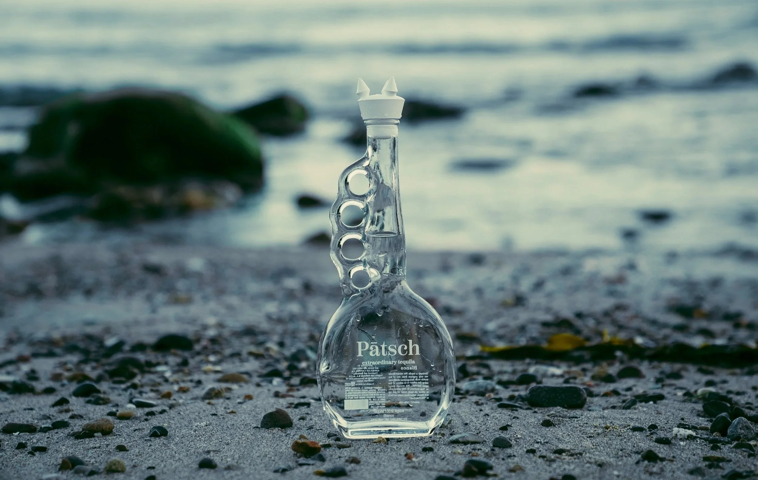 A clear glass bottle of Pátsch tequila standing upright on a pebbled beach with rocks in the background near the water's edge.
