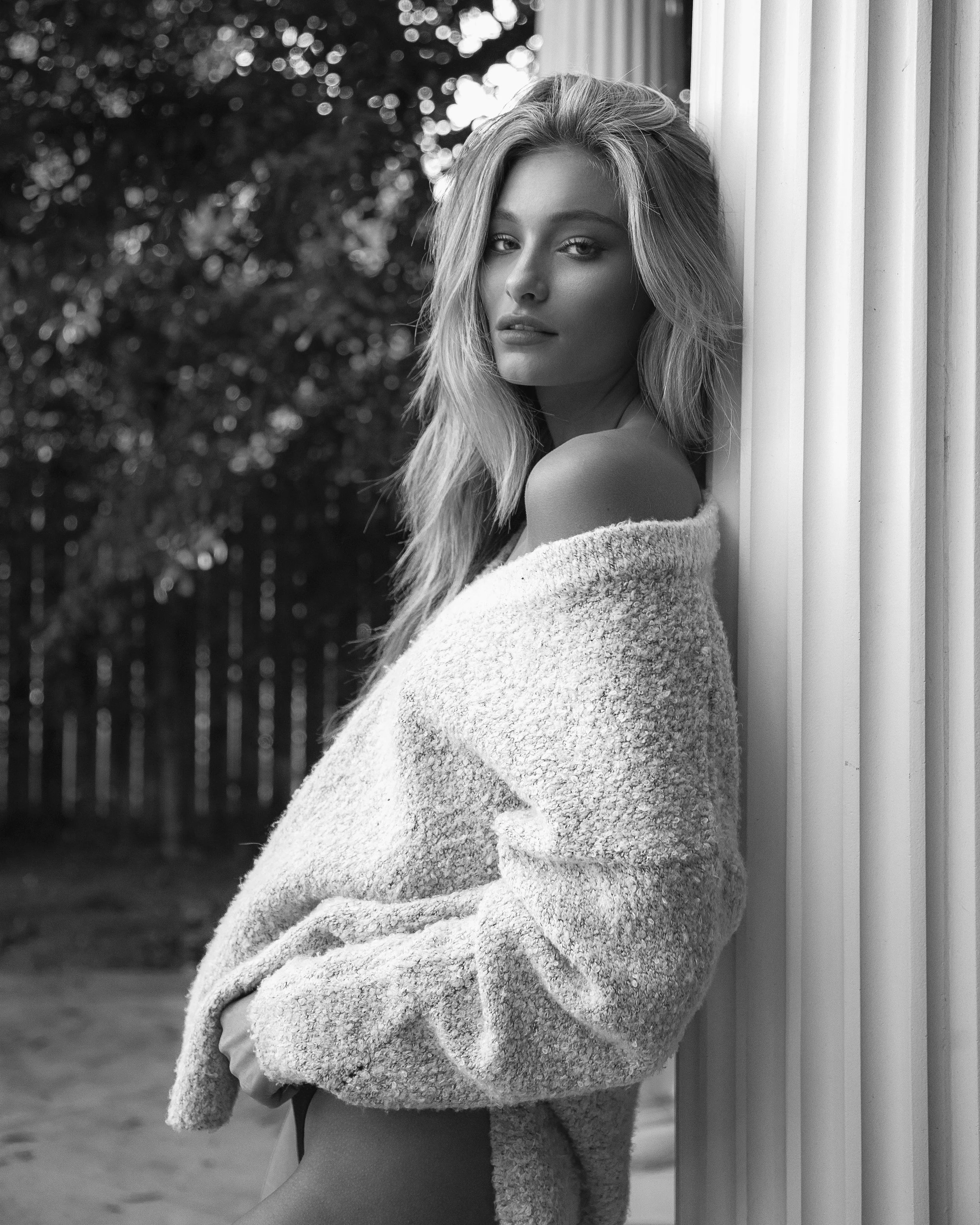 A black and white photograph of a young woman with long wavy hair leaning against a column outdoors, wearing a cozy off-the-shoulder sweater that exposes her shoulder, with trees and a fence in the background.