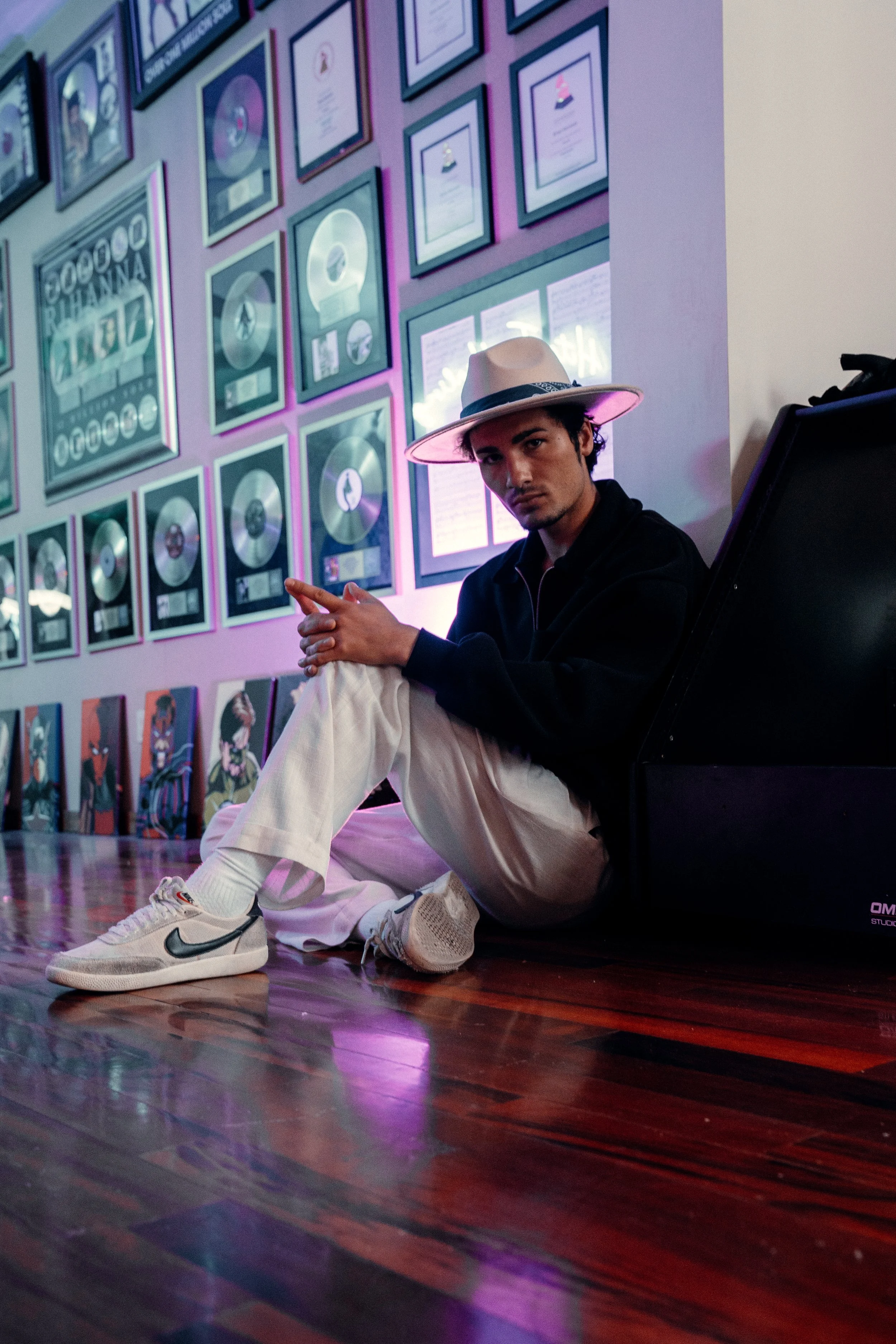 A young man sitting on the floor of a music studio or gallery, wearing a wide-brimmed hat, a dark jacket, and white pants, with a wall of gold and platinum record awards behind him.