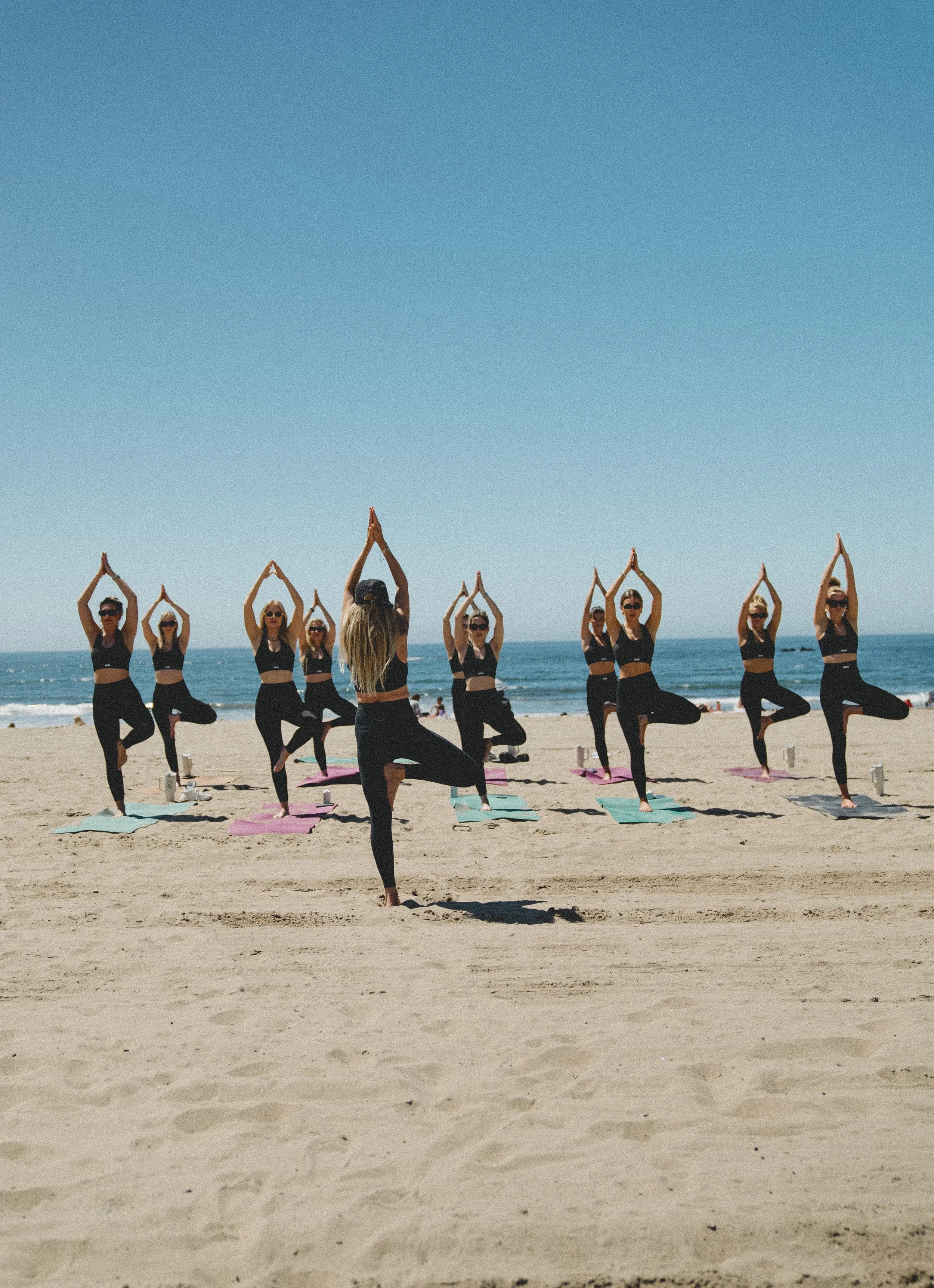 A group of women practicing yoga on the beach, standing on yoga mats with their hands raised above their heads and one leg bent at the knee, facing the ocean under a clear blue sky.