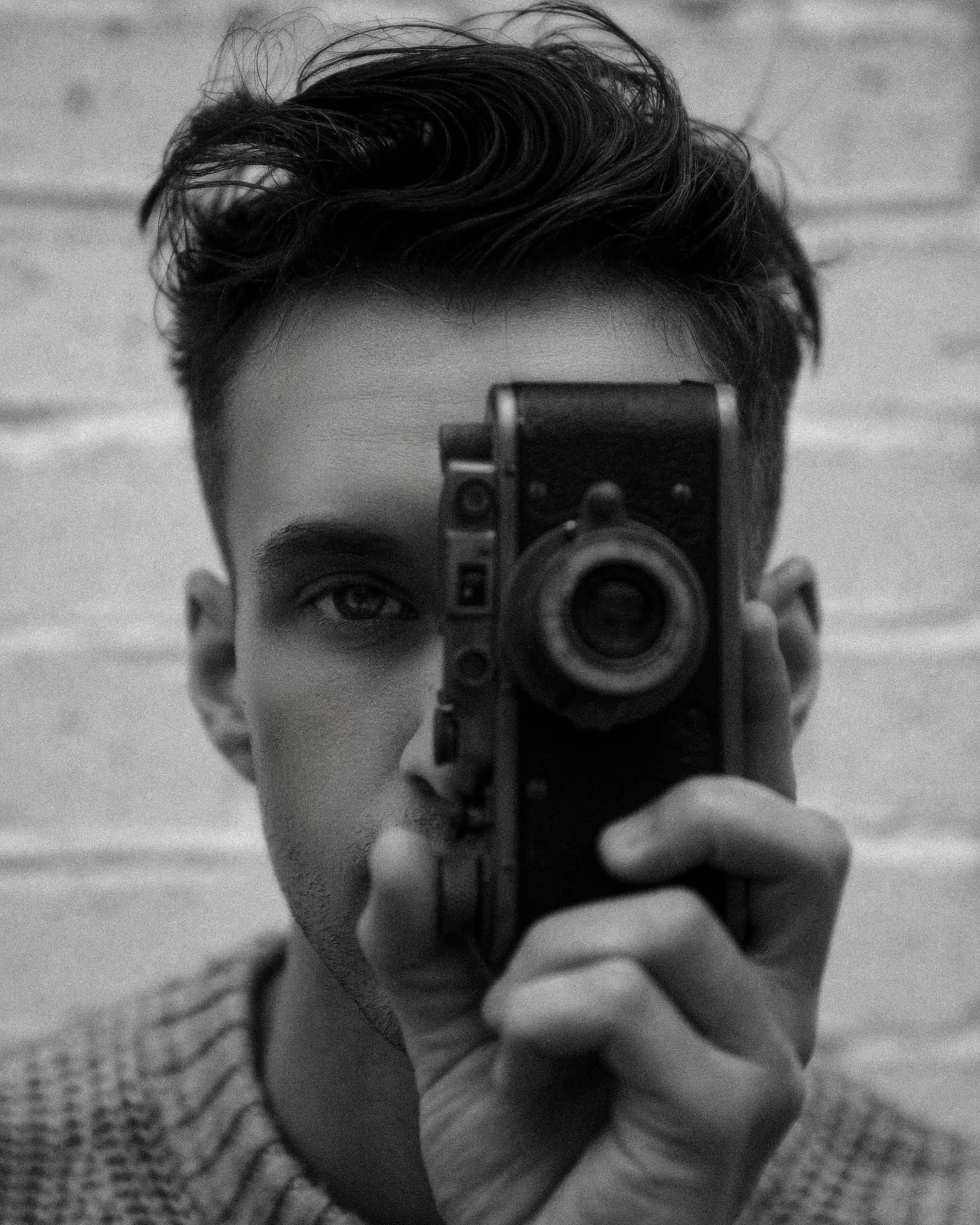 A young man with dark, styled hair taking a selfie with a vintage camera, partially obscuring his face, against a blurred background.