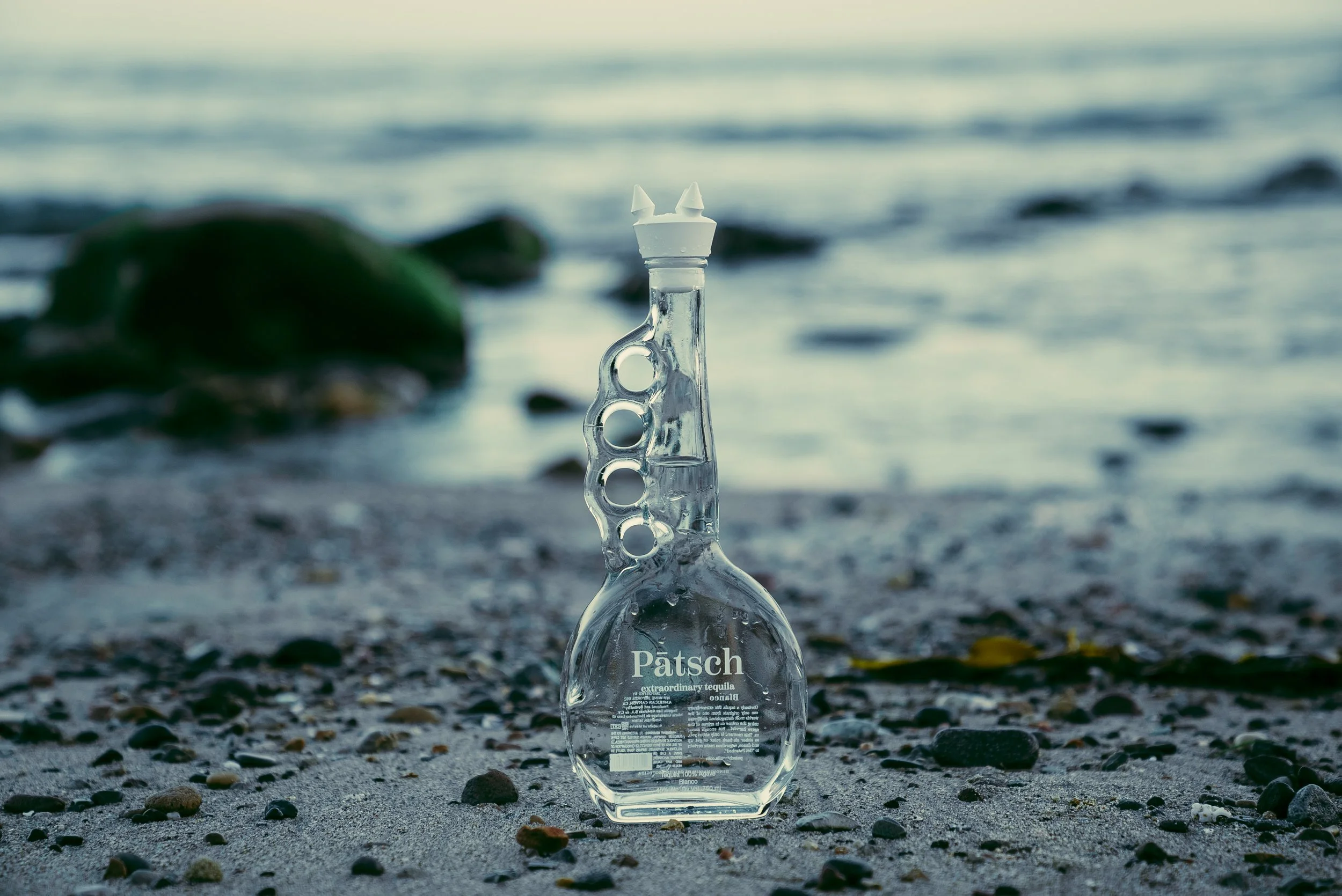 A clear glass bottle with the label 'Patsch extra ordinary tequila' placed upright on a sandy beach with small pebbles, near the water, with rocks and seaweed in the background.