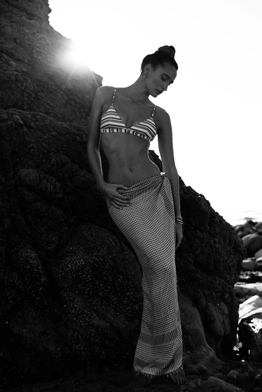 Black and white photo of a woman standing on rocks near the ocean, wearing a striped bikini top and patterned wide-leg pants, with the sun setting behind her.