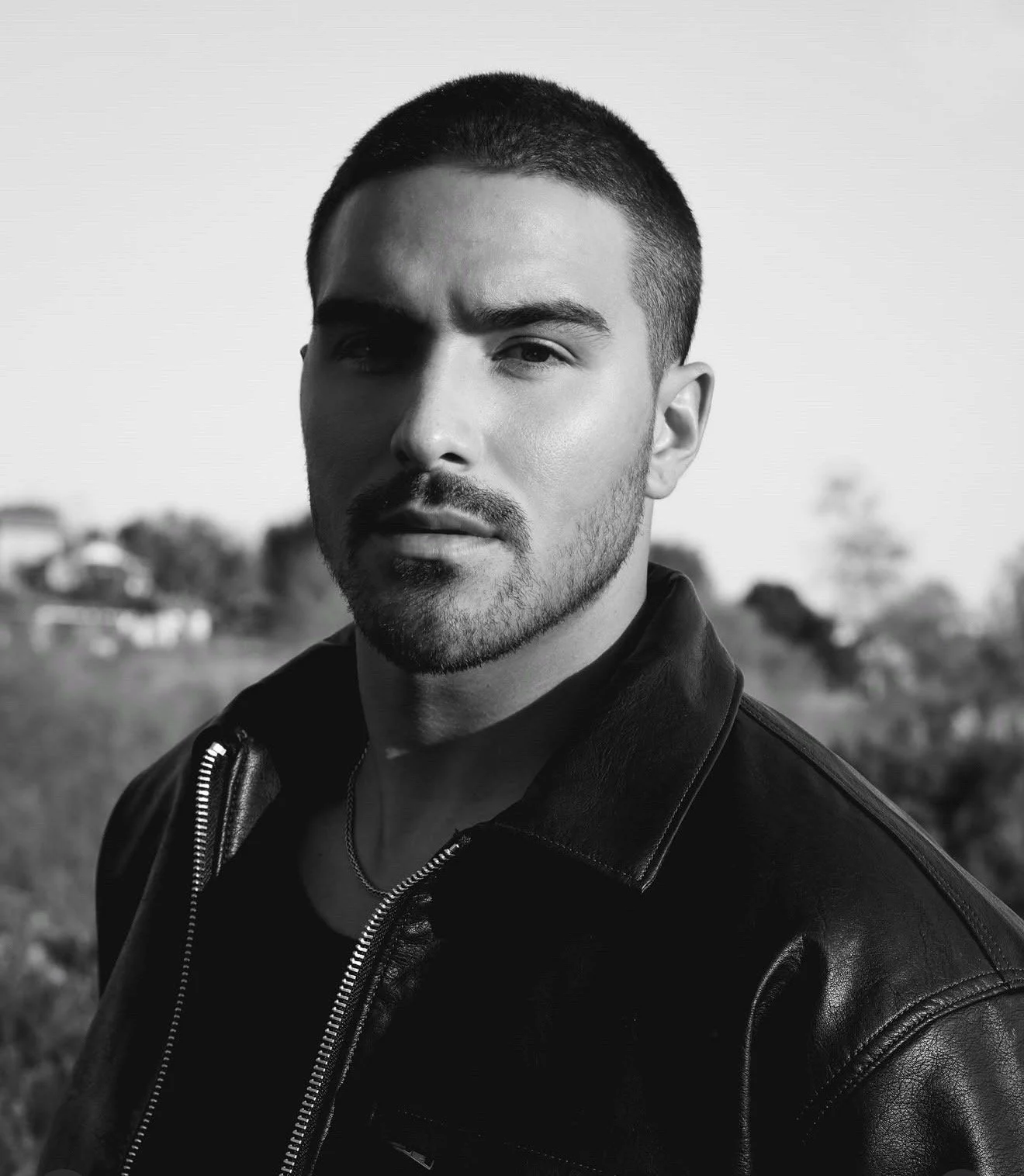 Black and white portrait of a young man with short hair, a beard, and wearing a leather jacket, outdoor background.