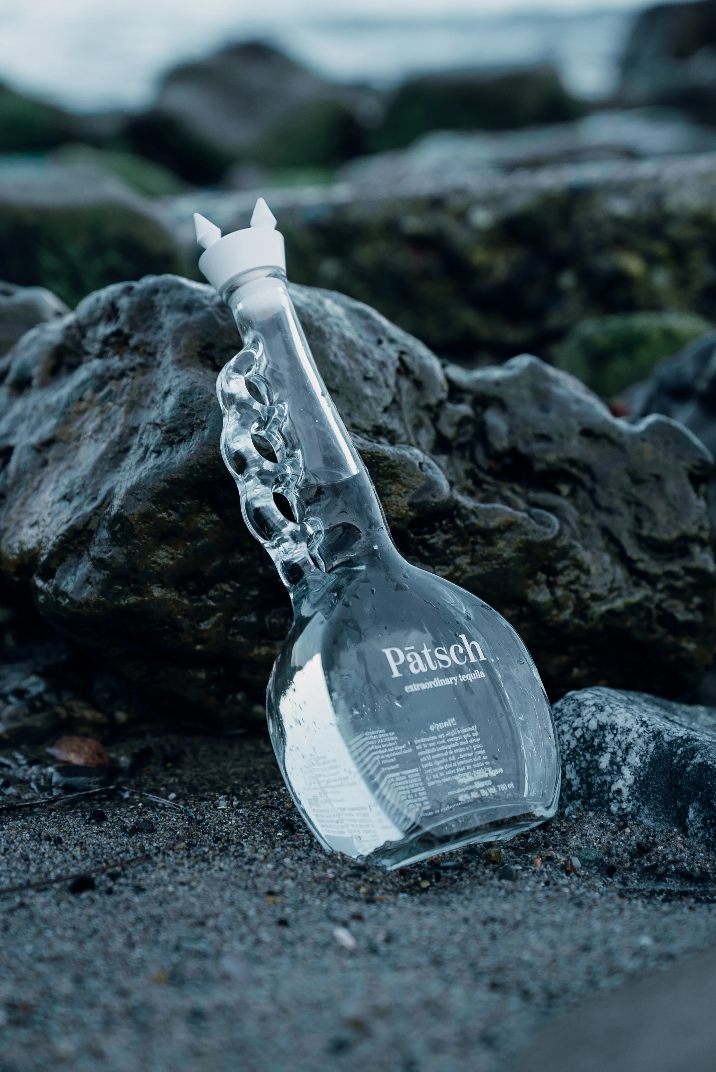 A glass bottle labeled 'Pätsch extraordinary tequila' lies on sandy ground among rocks near a body of water.