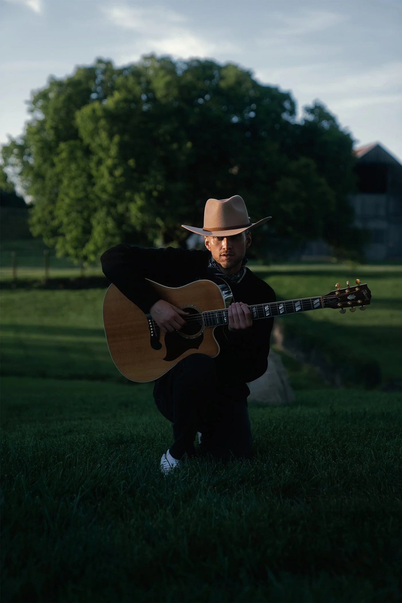 A man wearing a beige cowboy hat, black jacket, and white shoes, kneeling on the grass and playing an acoustic guitar outdoors with a large green tree in the background.