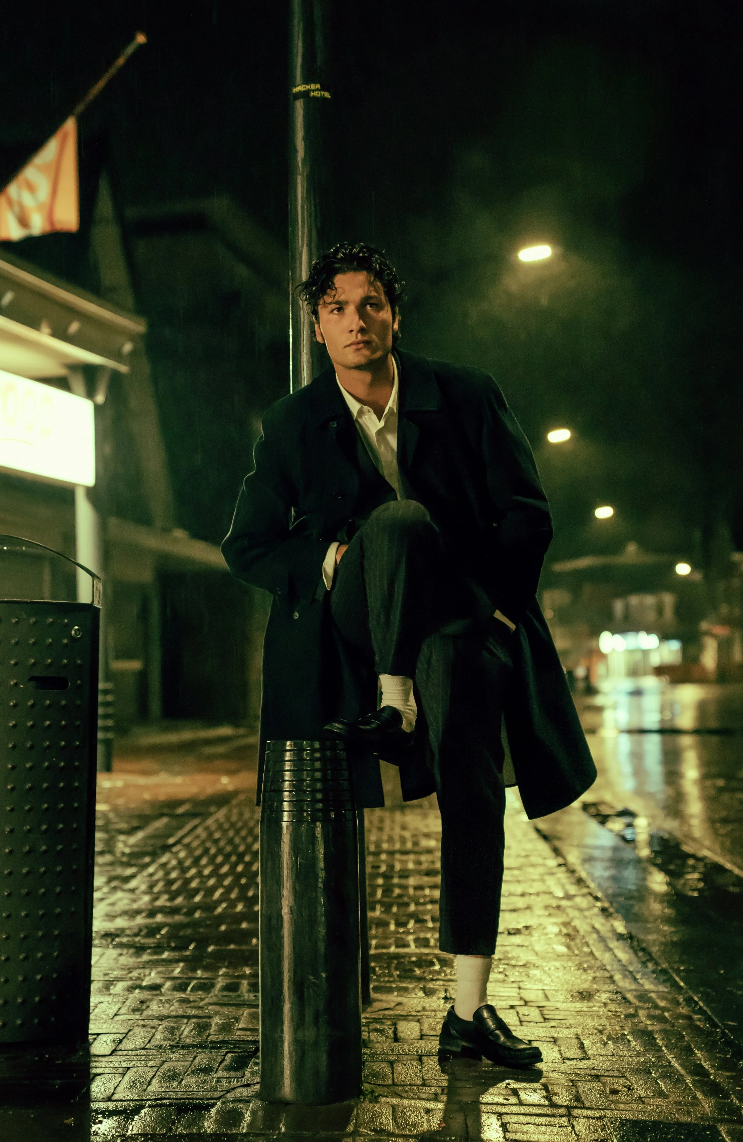 A man standing on a rainy city street at night, wearing a dark coat, with one foot on a metal post, and looking into the camera.