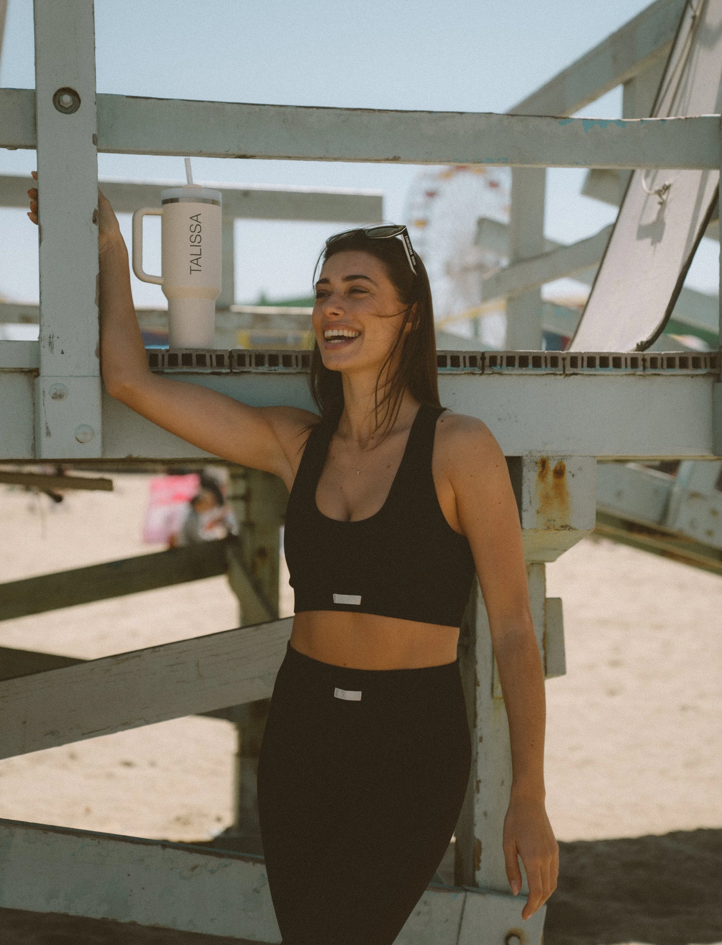 Woman in black workout clothing leaning against a lifeguard stand at the beach, smiling, with a large white tumbler labeled 'TALISSA' nearby.