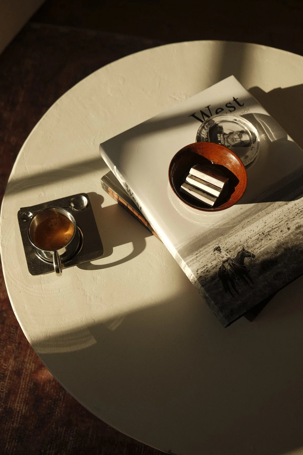 A white round table with a small espresso coffee cup on a metal saucer, a stack of magazines including one titled "West", and a round wooden container holding various metal objects. Sunlight creates shadows on the table.