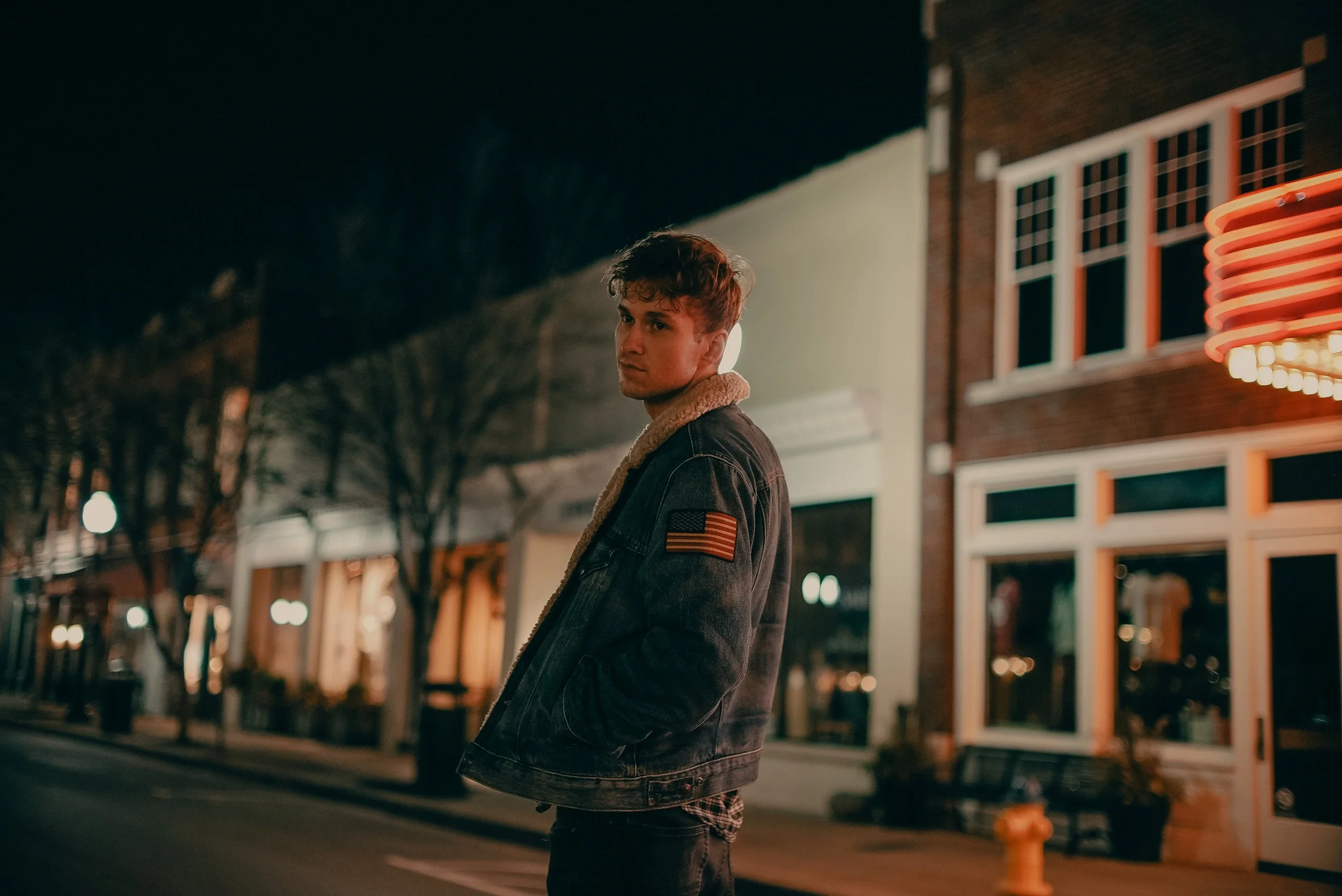 A young man standing on a city street at night, wearing a denim jacket with an American flag patch, looking to the side with hands in pockets, illuminated by street and building lights.