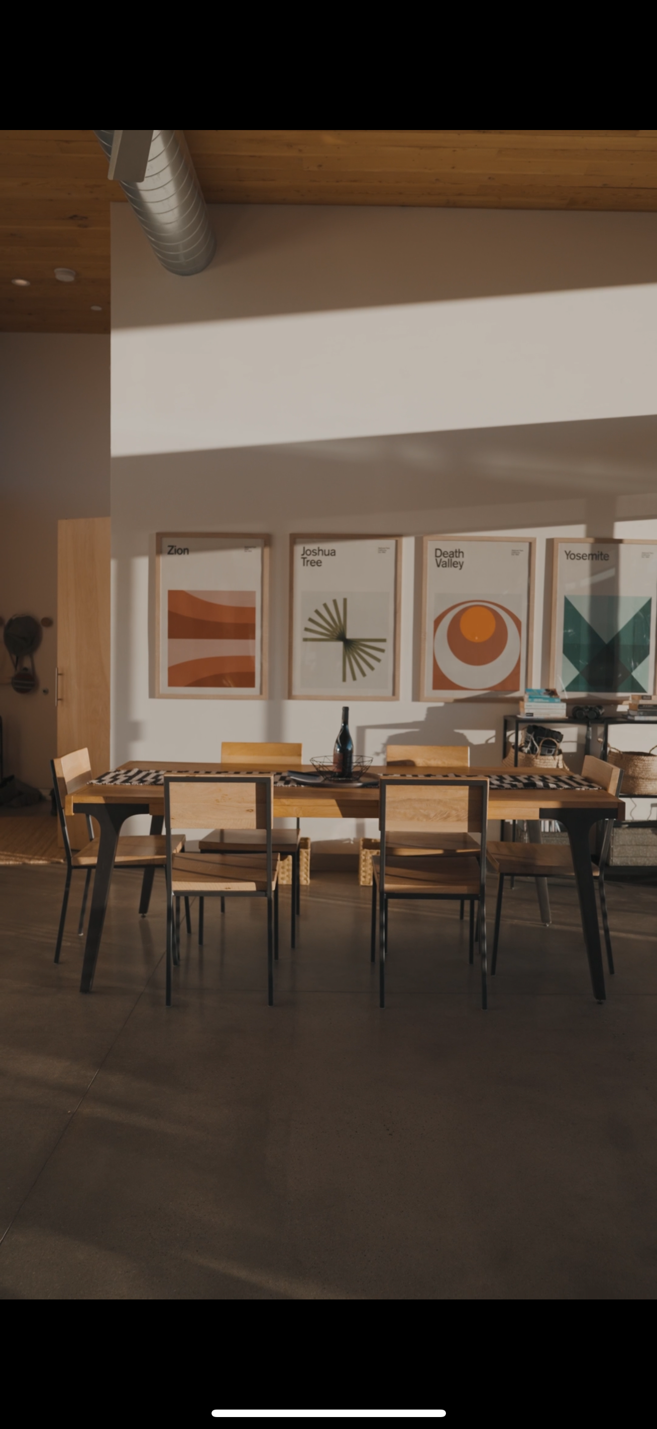 A dining room with a wooden table and six matching chairs. On the table is a bottle and a tray. The wall behind has four framed artwork with abstract designs labeled Zion, Joshua Tree, Death Valley, and Yosemite. To the right, a black shelf holds boo