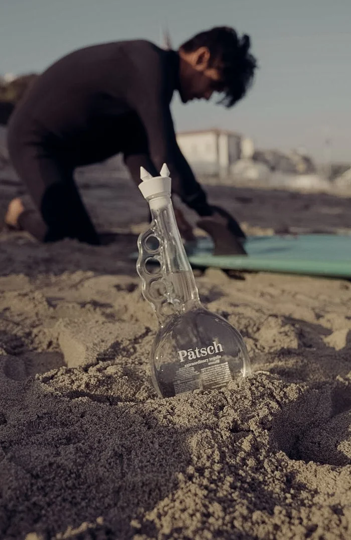 A glass bottle of Patsch brand alcohol partially buried in the sand on a beach with a person in the background in a black outfit preparing to surf or swim.