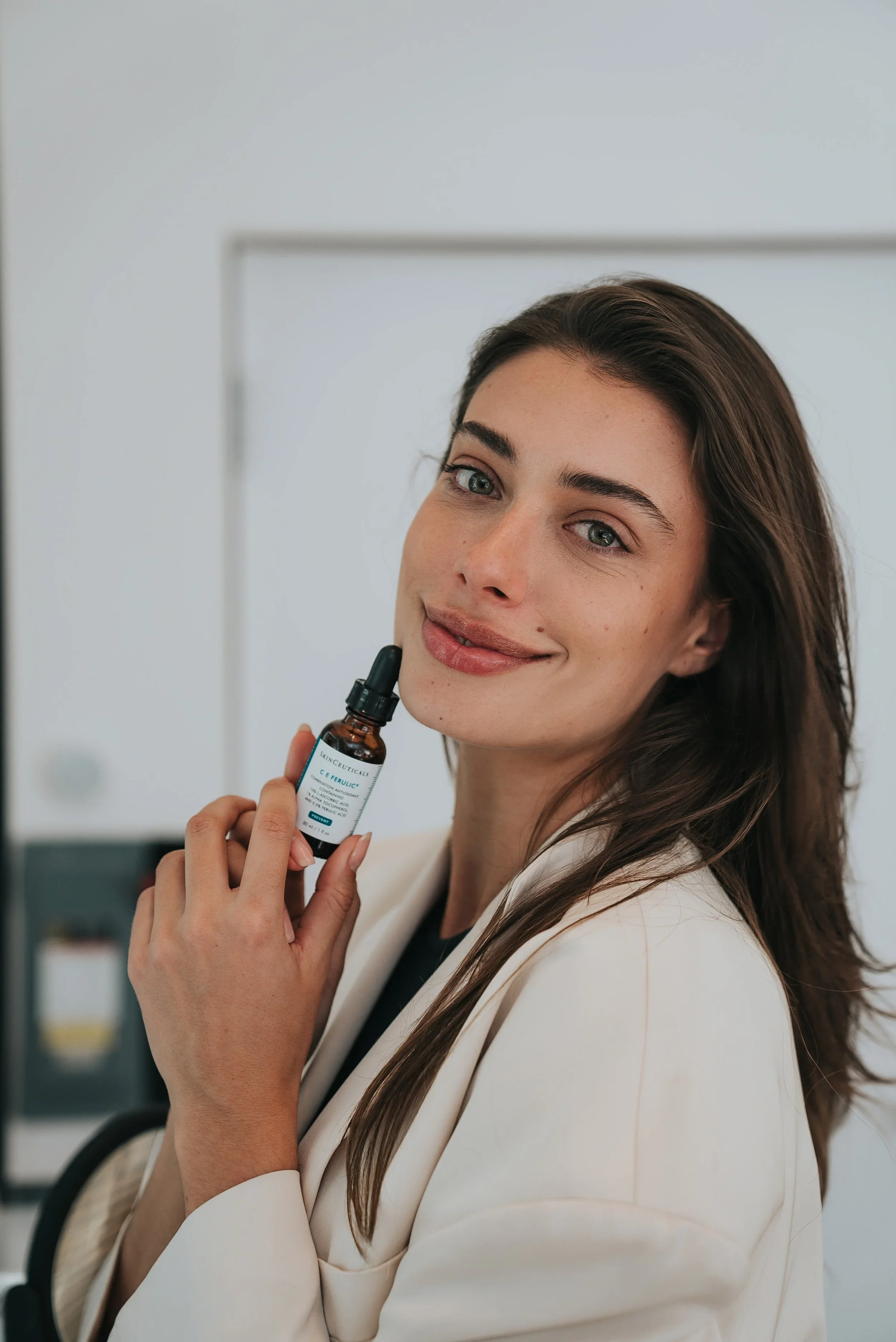 A woman with long brown hair holding a small brown bottle with a dropper, looking at the camera and smiling, in a bright white room.