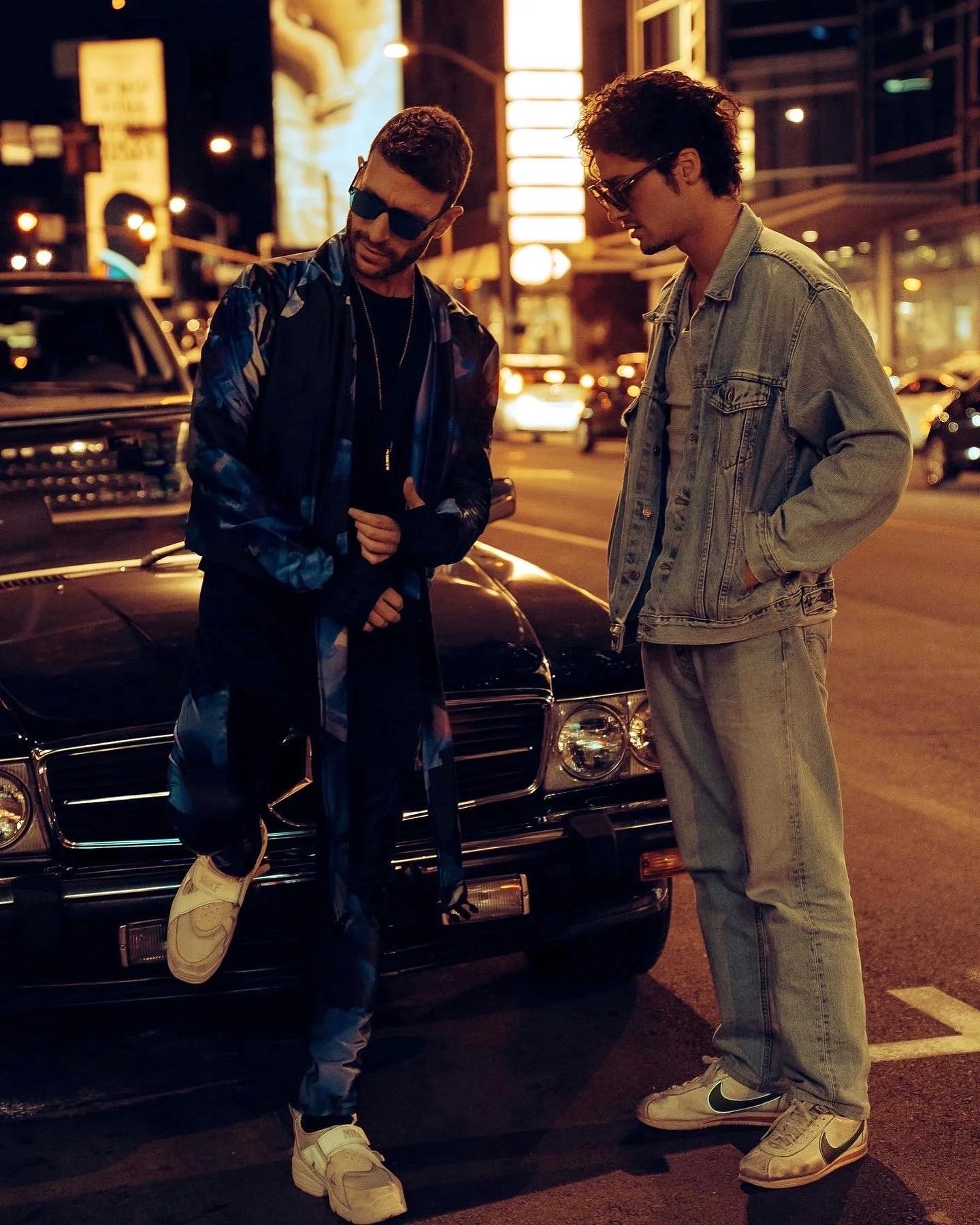 Two young men with casual streetwear stand next to a black vintage Mercedes-Benz in a city street at night, illuminated by streetlights and neon signs.