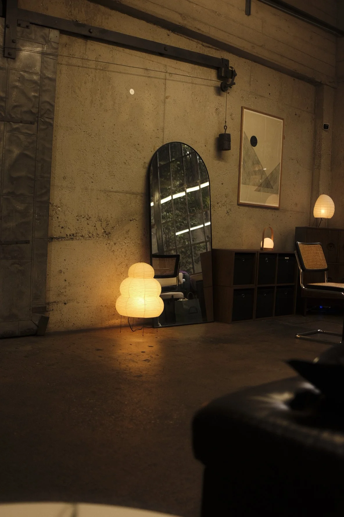 Interior of a room with a large arched mirror leaning against a concrete wall, lit by soft warm lighting including paper lantern lamps and a small table lamp, with modern abstract wall art, a wooden shelving unit, and a chair.
