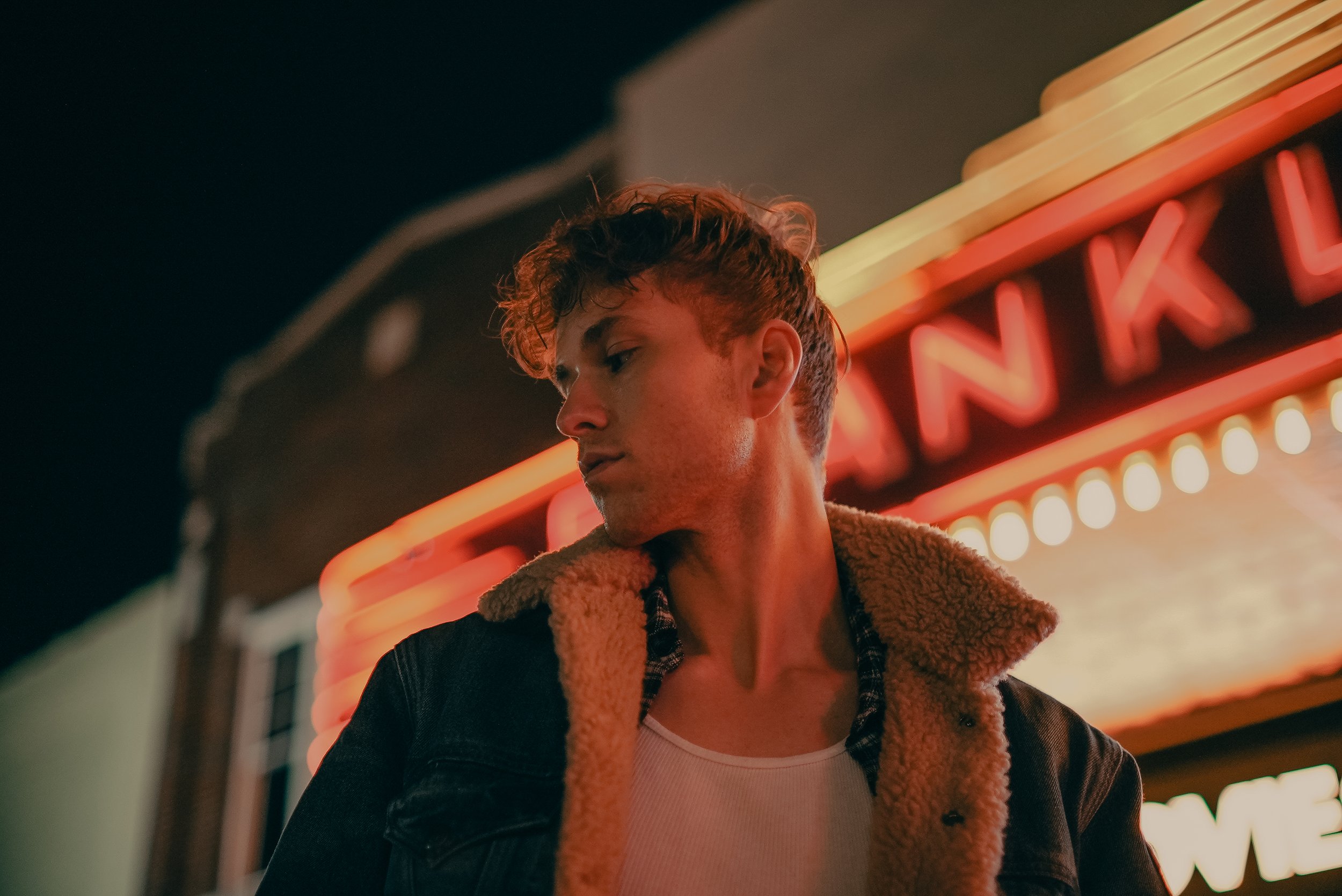 Young man with curly hair and a denim jacket standing outside at night near a neon-lit sign.
