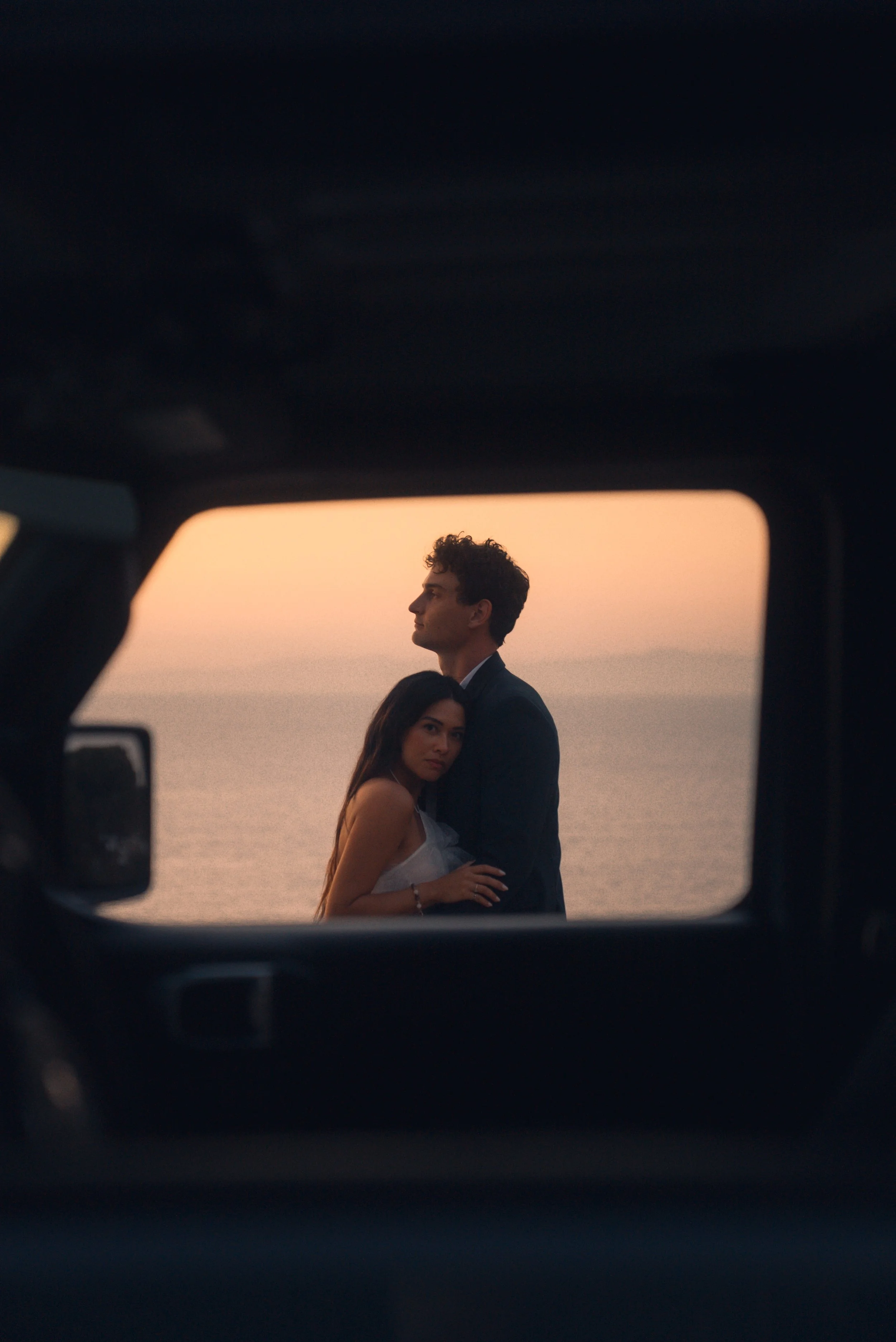 A couple is seen through the window of a vehicle at sunset, with the ocean in the background. The woman is leaning on the man's chest, both gazing thoughtfully into the distance.