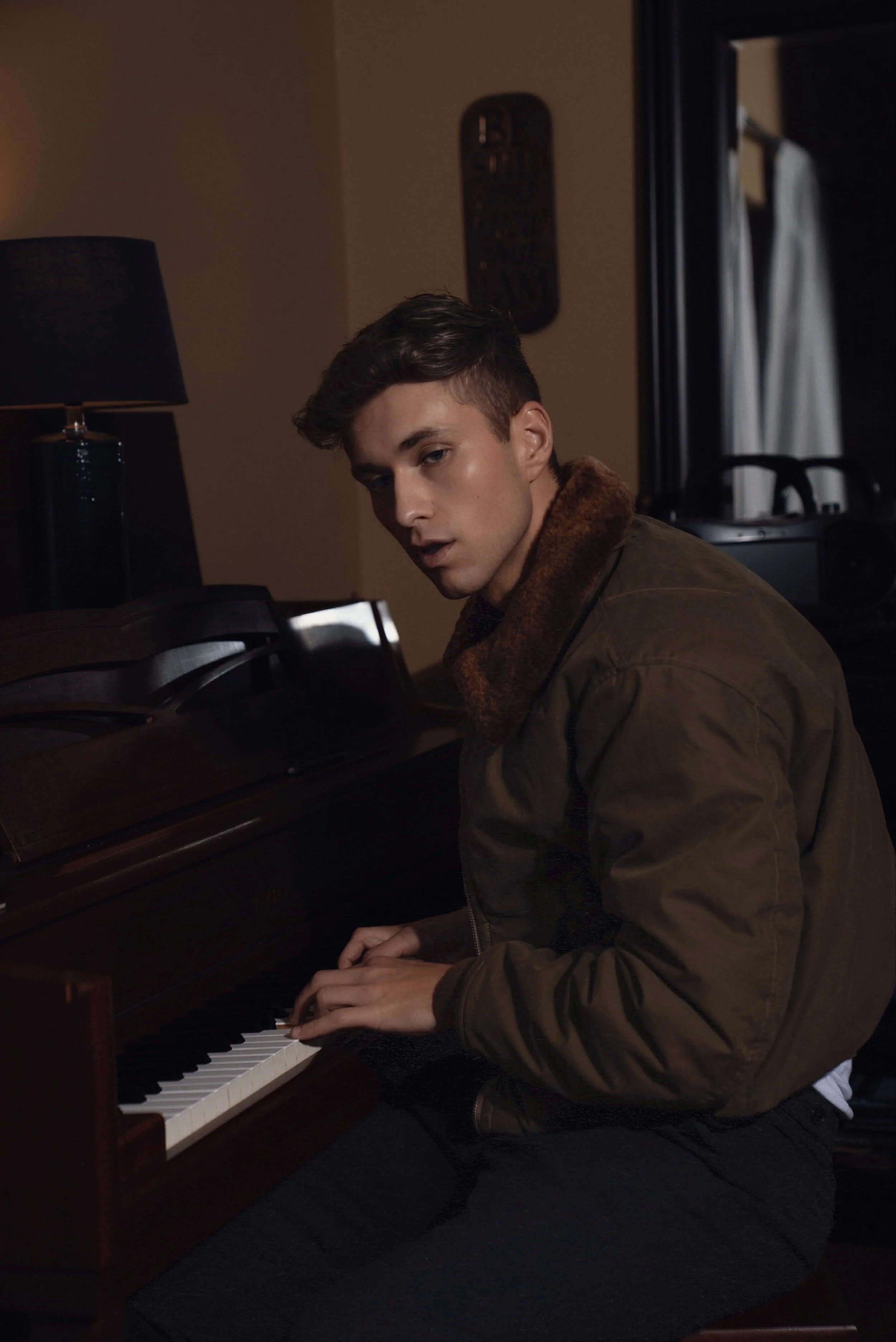 Young man sitting at a piano, looking at the camera, wearing a brown jacket with a fur collar, in a dimly lit room.