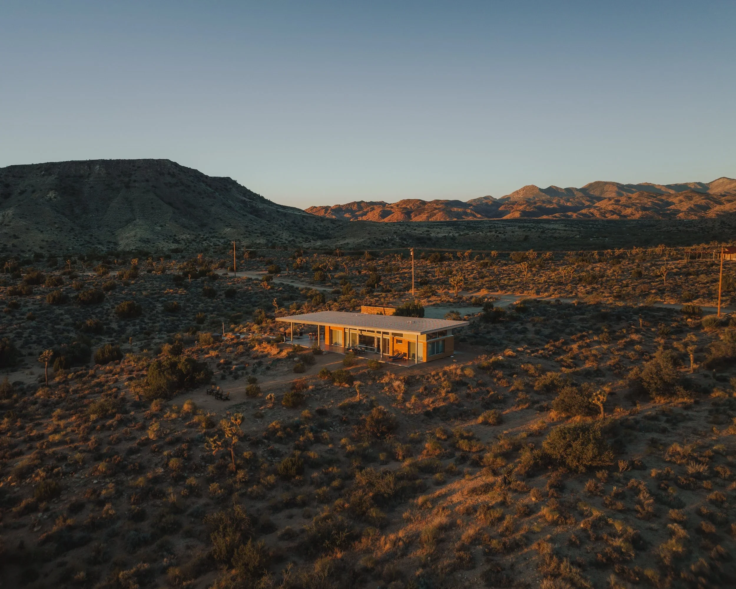 A modern house with large glass windows and a flat roof set in a desert landscape at sunset, surrounded by sparse bushes and distant mountains.