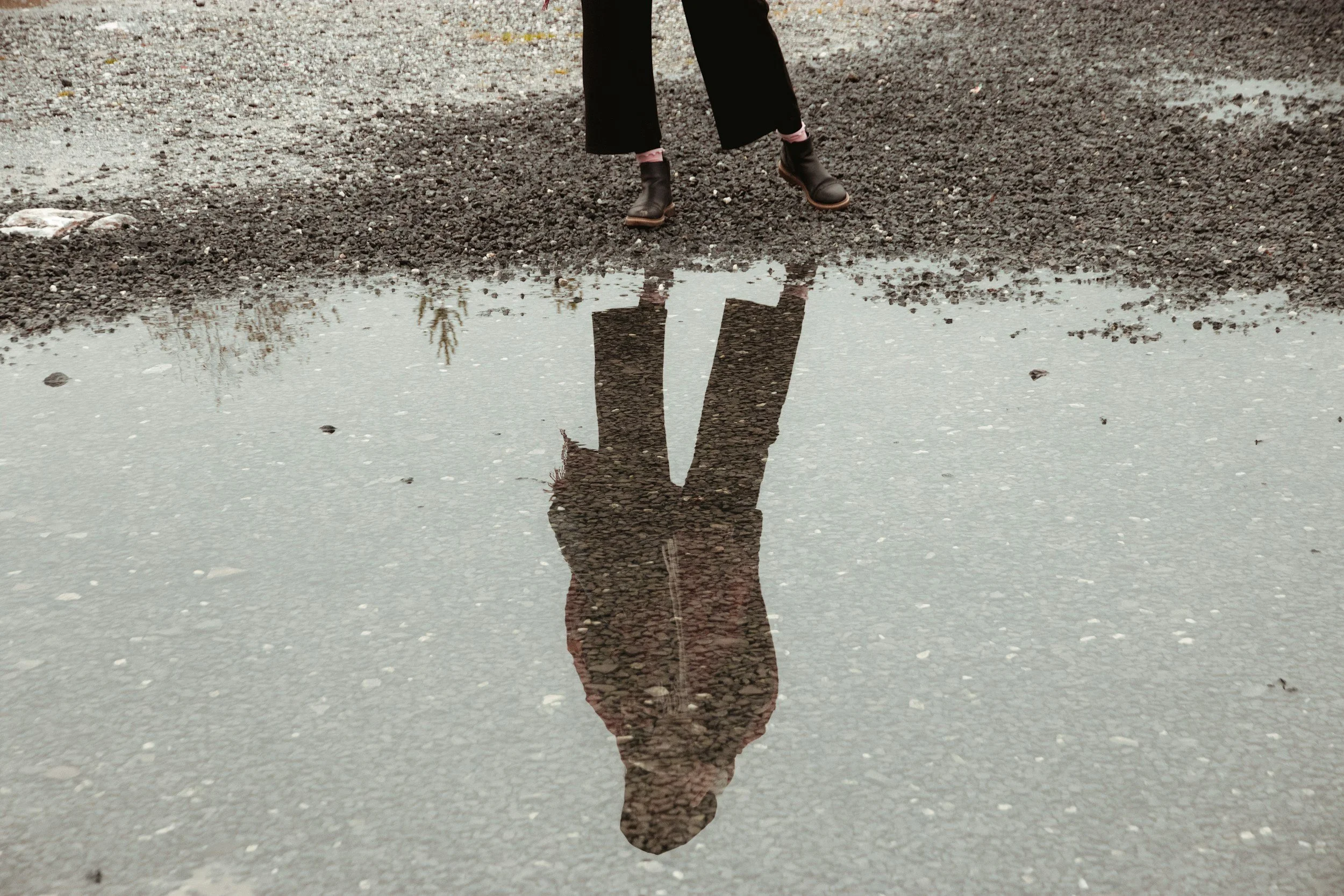 Person standing on gravel ground, reflected in a puddle of water.