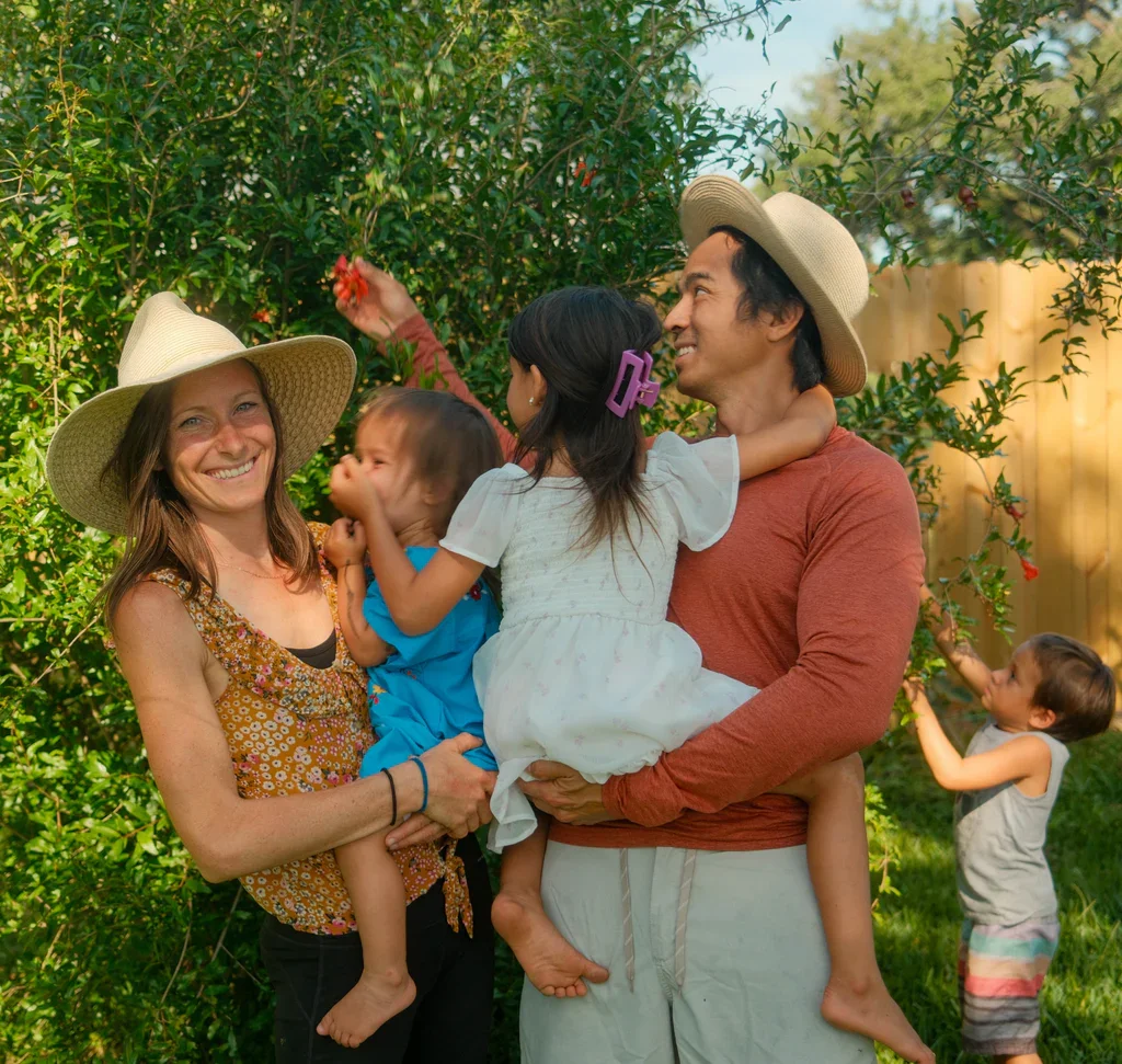 Family of four enjoying a sunny day in a garden, with two children and two adults wearing sun hats, surrounded by greenery.