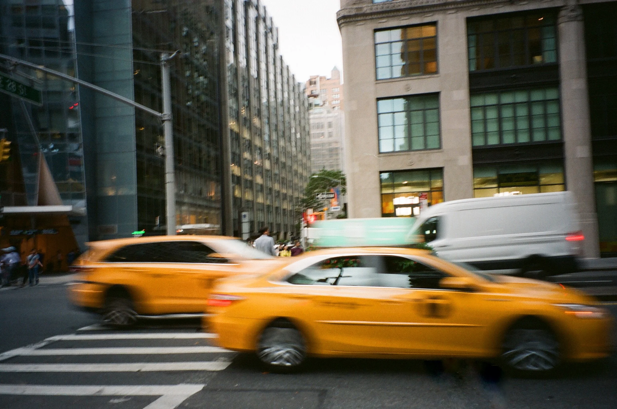 Blurred yellow taxi cab and white delivery van on city street with tall buildings in the background.