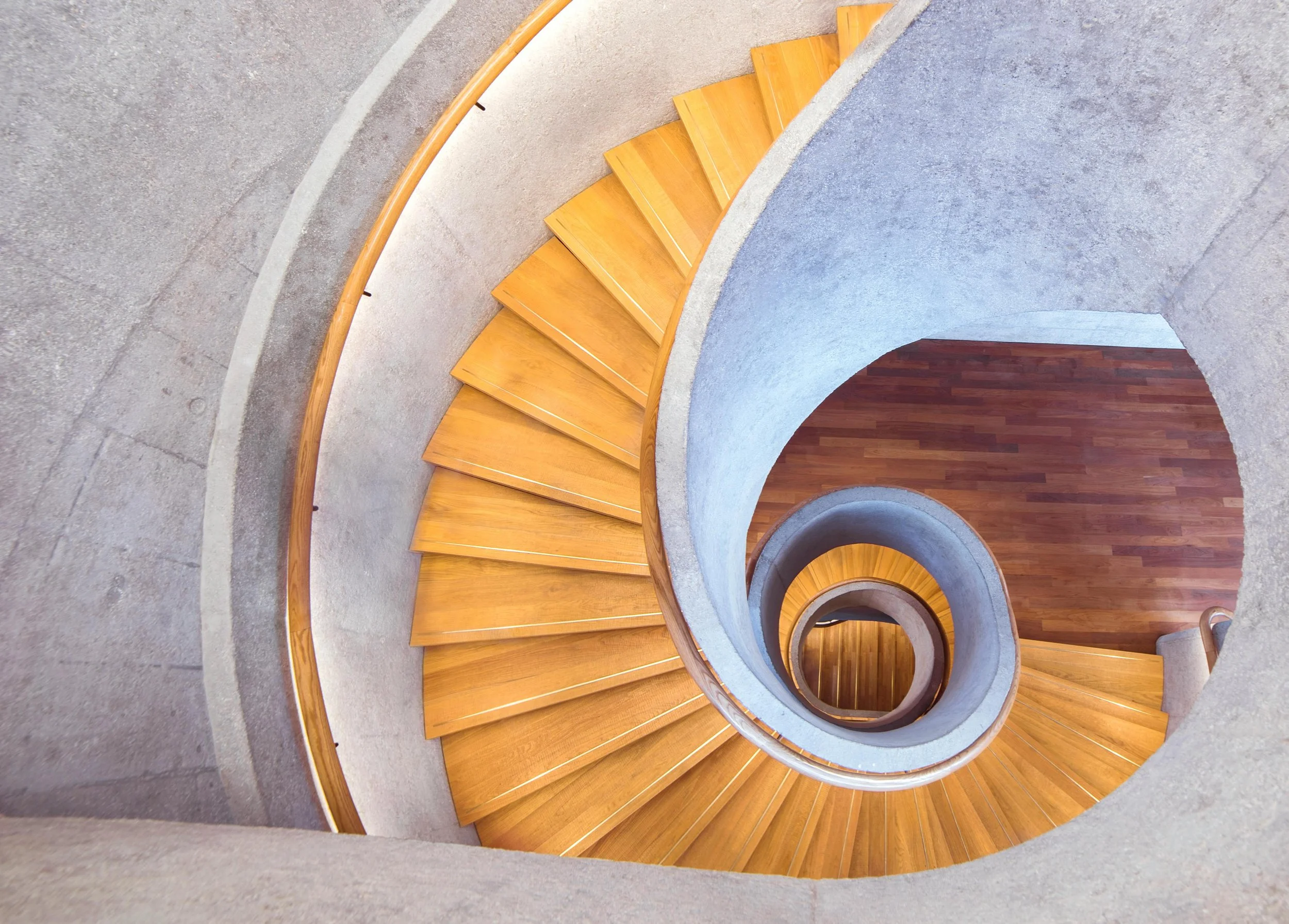 Top-down view of a spiral staircase with wooden steps and a concrete railing, leading downward to a wooden floor.