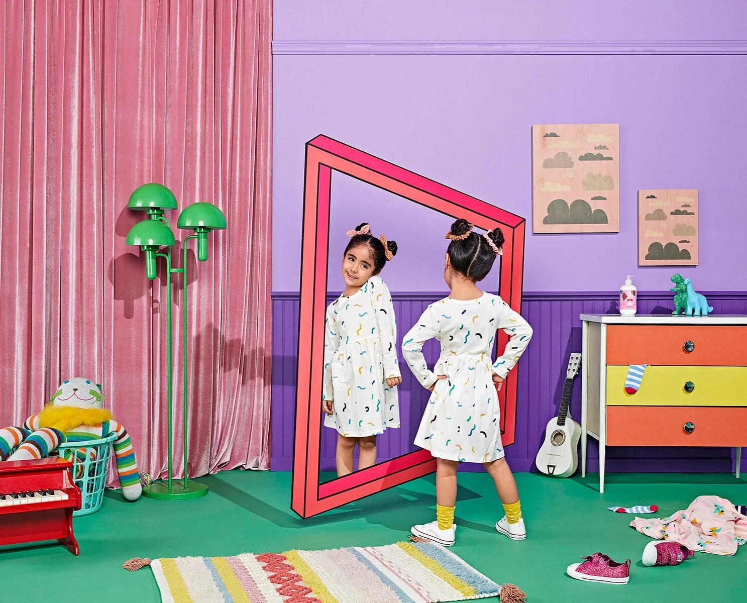 Two girls are standing in a colorful room with purple walls. One girl is looking into a large mirror and smiling, while the other girl is standing with her back to the mirror. The room has pink curtains, a green floor lamp, a rainbow-colored rug, a d