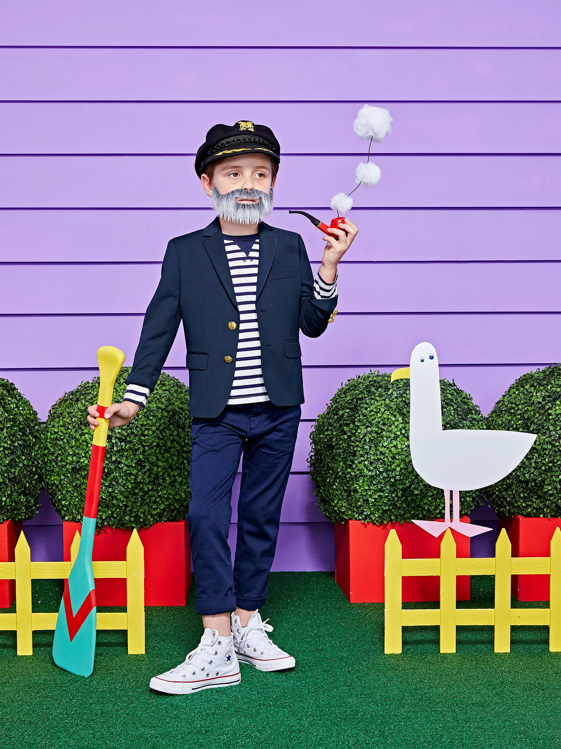 Child dressed as a sailor with a dark blazer, striped shirt, and white sneakers, holding a colorful paddle and smoking a pipe with cotton ball smoke. The kid has a faux gray beard and is standing outdoors with purple-paneled wall, green bushes, a whi
