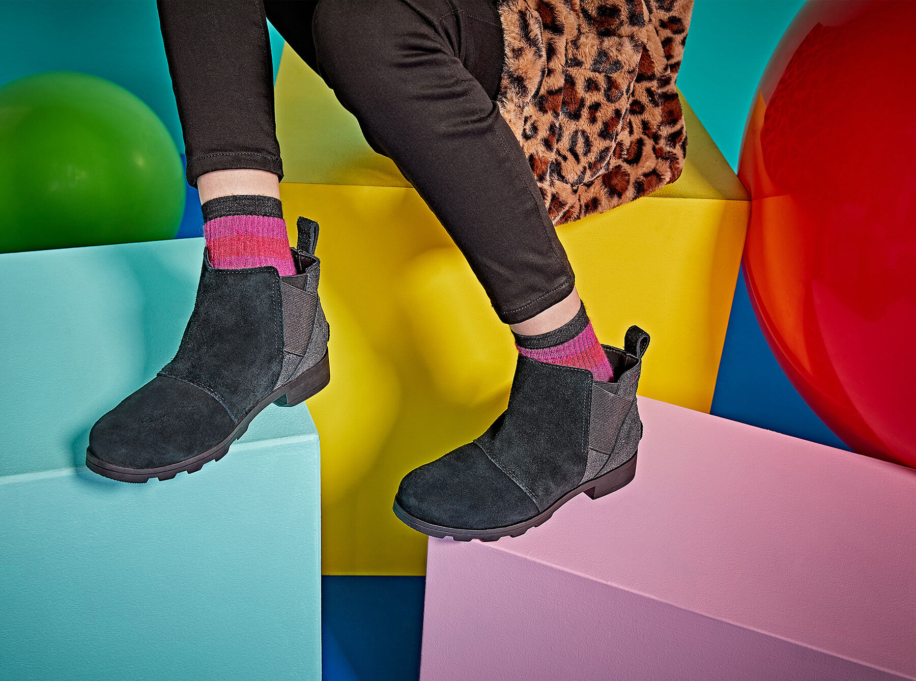 Close-up of a person sitting on multicolored block-shaped seats in a playful, colorful setting. The person is wearing black boots, ripped black pants, and colorful socks with pink on the top and black at the bottom, with a leopard-print coat visible.