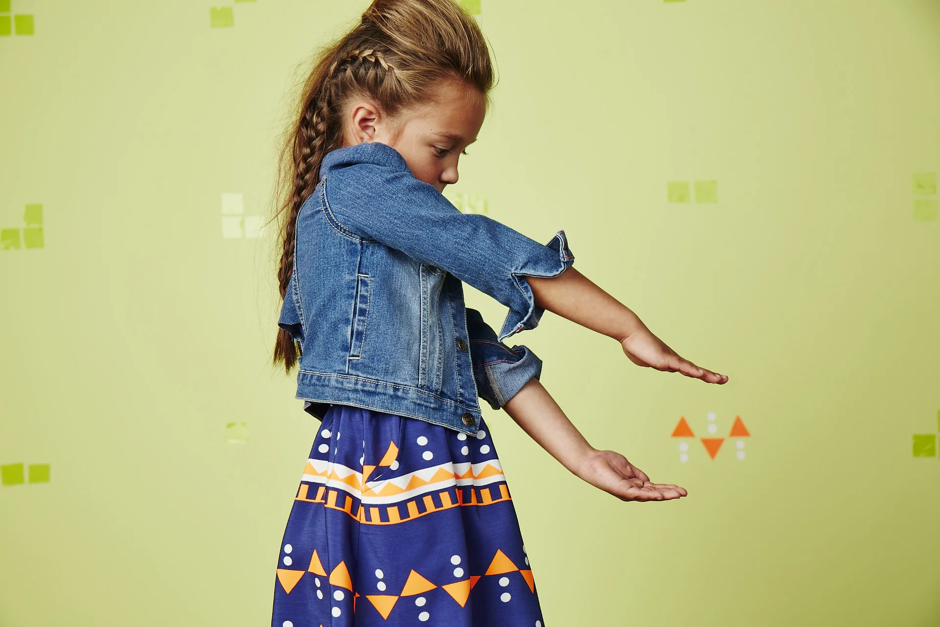 Young girl in denim jacket and patterned skirt, extending her hands outstretched in front of her against a light green background.