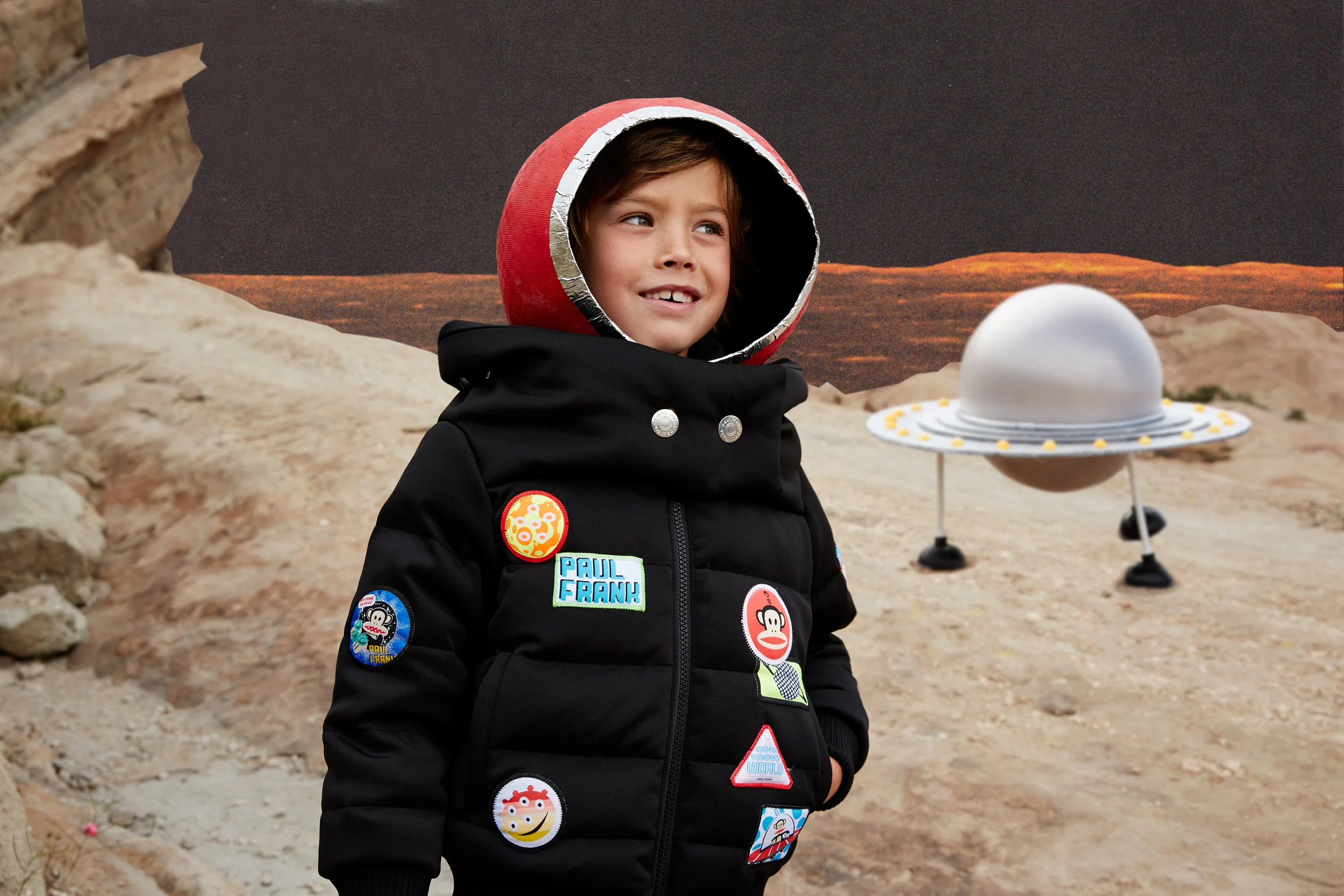 Child dressed as an astronaut with patches on a black jacket, wearing a helmet, standing in front of a space-themed backdrop with a UFO and planets.