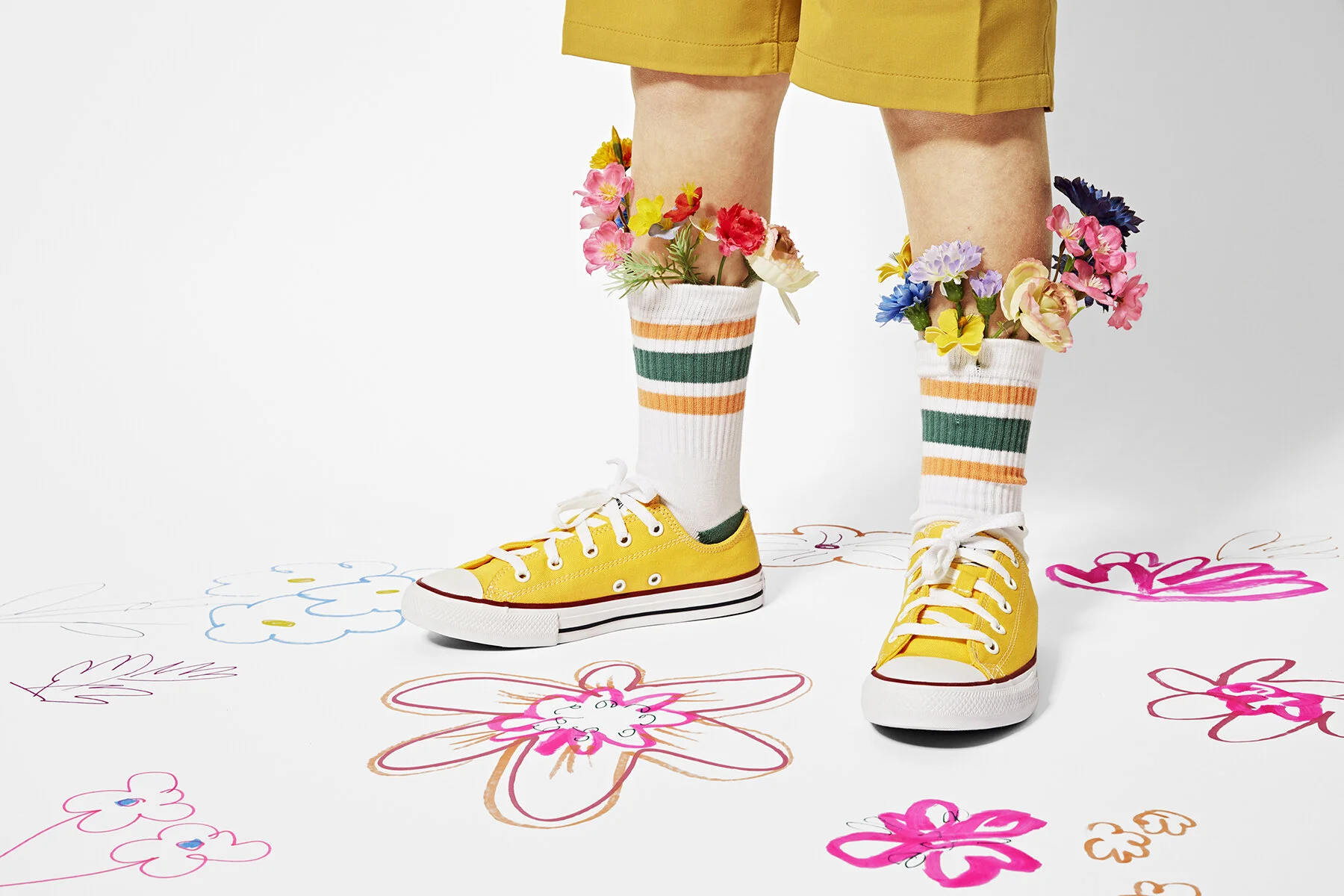 Person wearing yellow sneakers, white socks with orange and green stripes, and mustard-colored shorts, standing on a white surface decorated with colorful, childlike drawings of flowers and butterflies. The person's legs are stuffed with artificial f