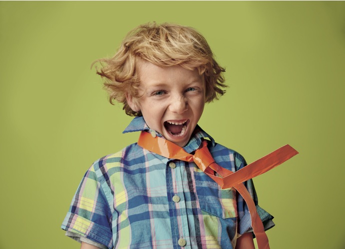 A young boy with curly blonde hair, wearing a colorful plaid shirt and an orange ribbon around his neck, shouting or yelling with an expressive look against a light green background.