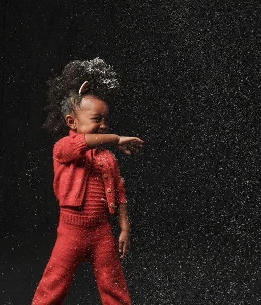 A young girl in red knit clothing playing with white powder, possibly flour or powder, against a black background.