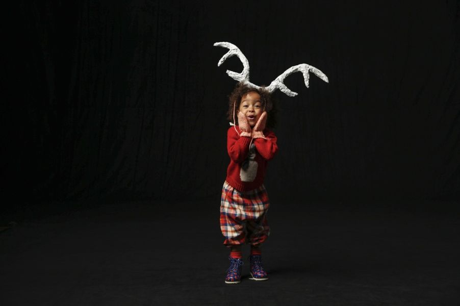 A young girl dressed in Christmas-themed clothing with plaid pants, red sweater, and reindeer antlers, standing against a black background, making a surprised face.