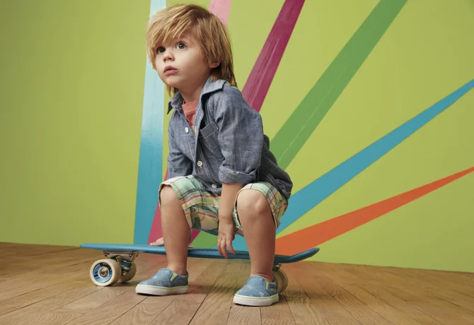 A young boy with messy light brown hair wearing a denim jacket and plaid shorts, sitting on a skateboard indoors against a colorful wall with diagonal stripes.