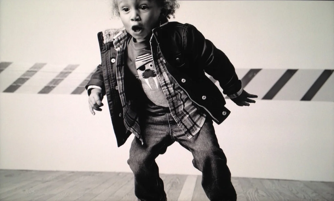 A young boy standing indoors against a wall with striped tape, wearing a jacket, plaid shirt, and jeans, with a surprised or curious expression.