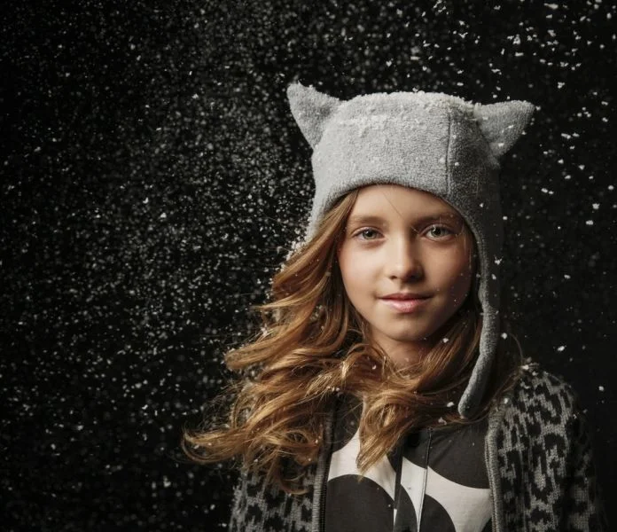 A girl with long, wavy hair wearing a gray hat with cat ears, standing against a black background with falling snow.