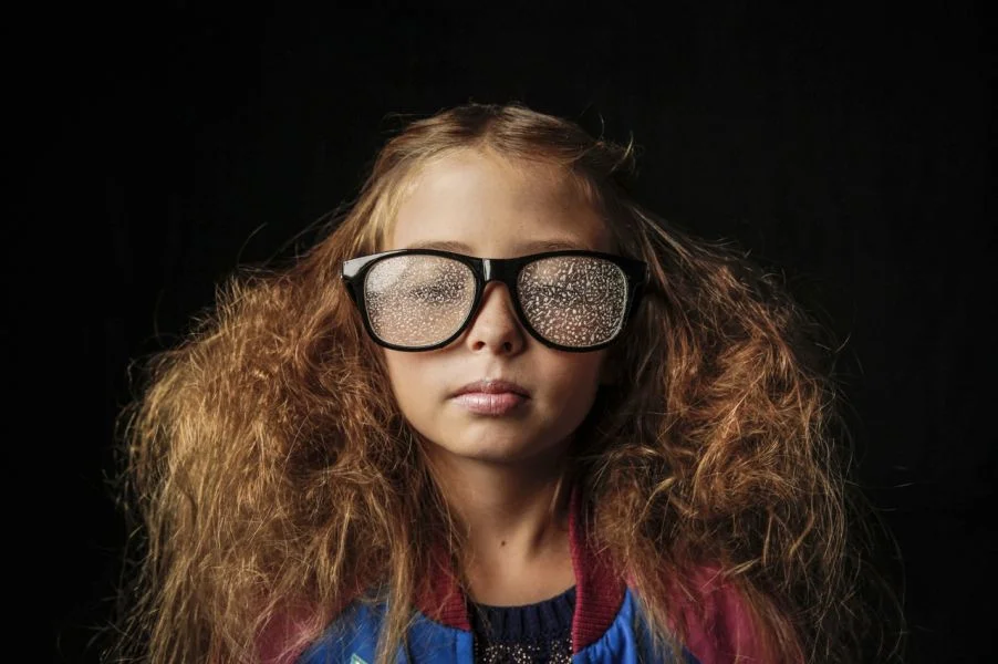 A young girl with large, curly red hair wearing oversized glasses with a raindrop-covered lens, against a black background.