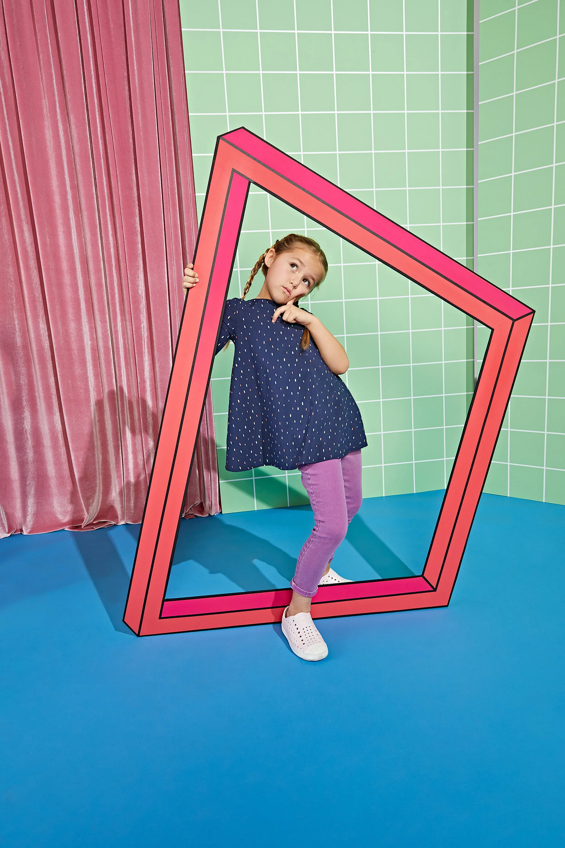 A young girl with braided hair, wearing a navy blue polka dot dress, purple pants, and white shoes, stands inside a large illustrated pink picture frame, holding the frame with one hand and touching her chin thoughtfully, in a colorful setting with g
