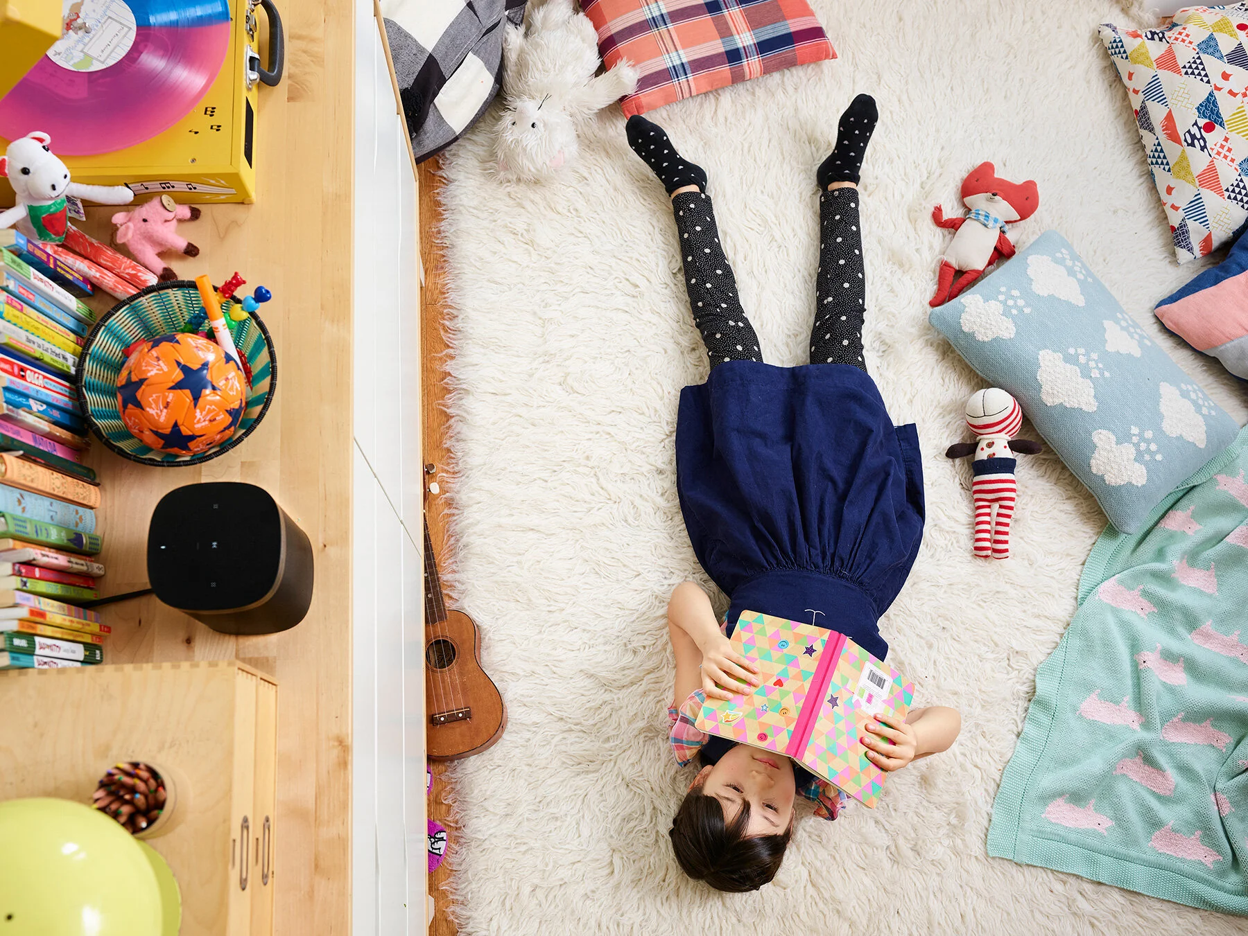 A girl lying on a plush white rug in a children's room, holding a colorful patterned book over her face, surrounded by toys, stuffed animals, and pillows.