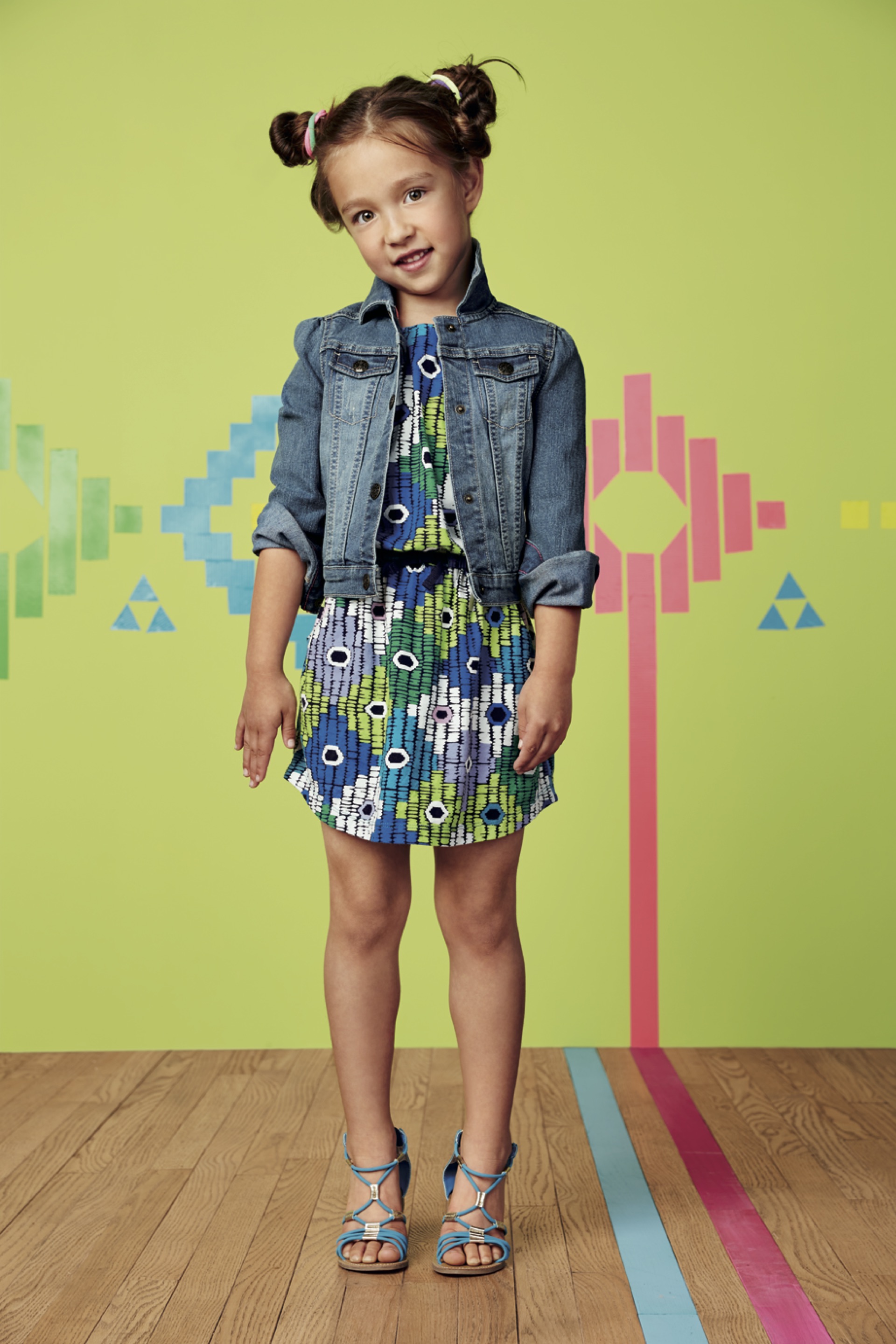 Young girl with colorful dress, denim jacket, and high-heeled sandals standing on a wooden floor with a vibrant, geometric, and video game-inspired background.